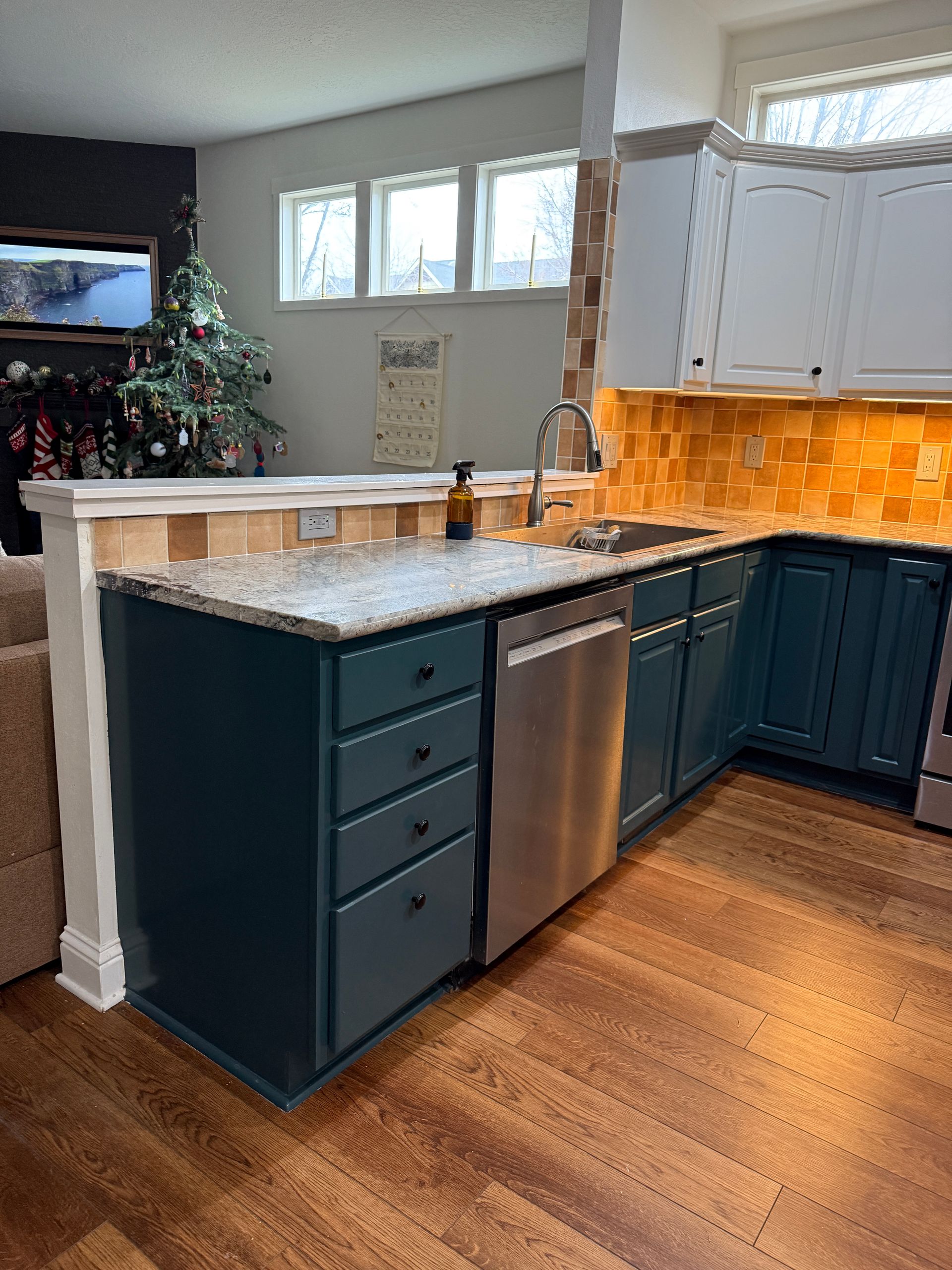 Modern kitchen with teal cabinets, granite island, stainless dishwasher, and warm orange backsplash