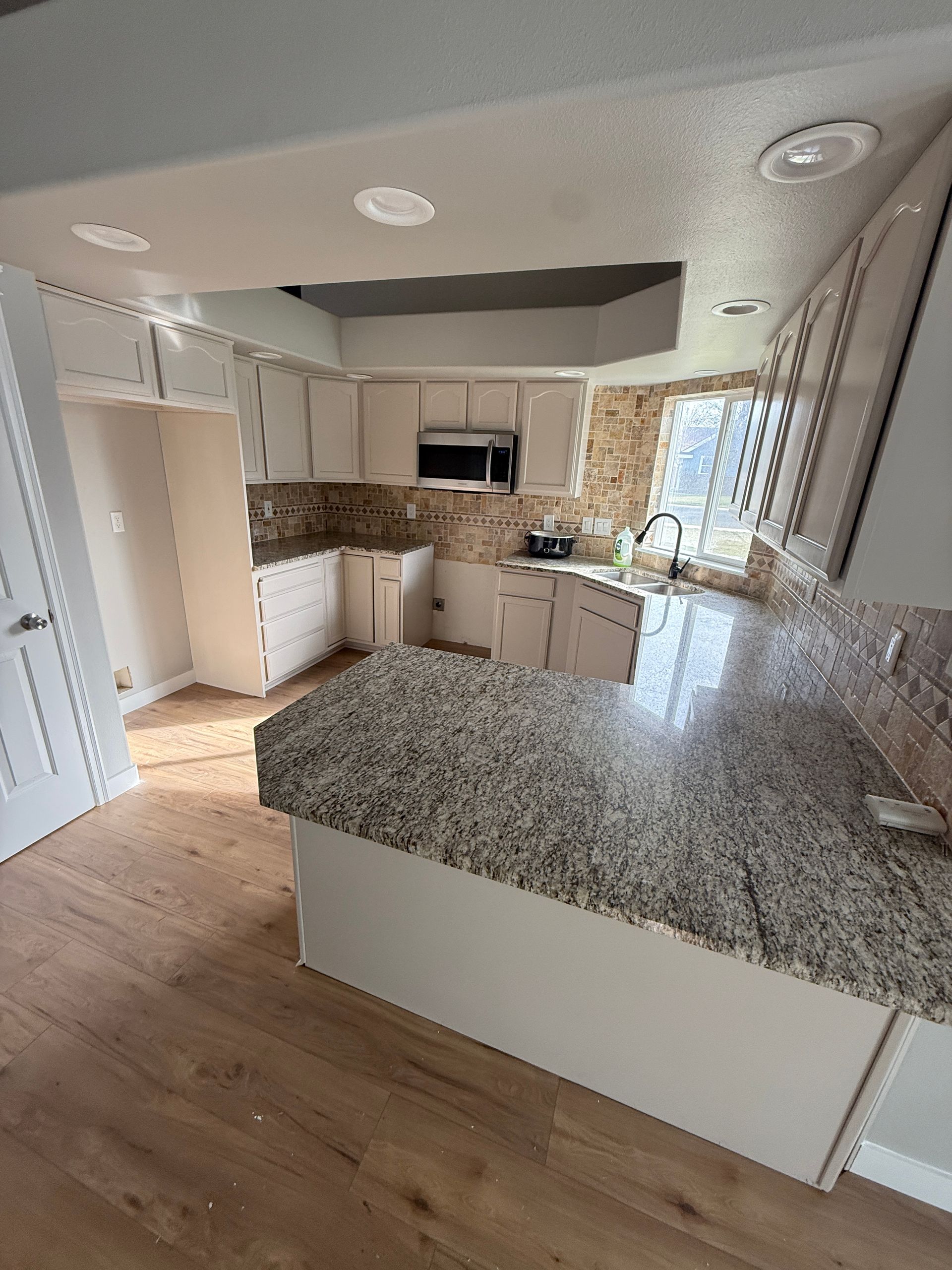 Bright kitchen with granite island, wood floors, white cabinets, and sunlight through a window.