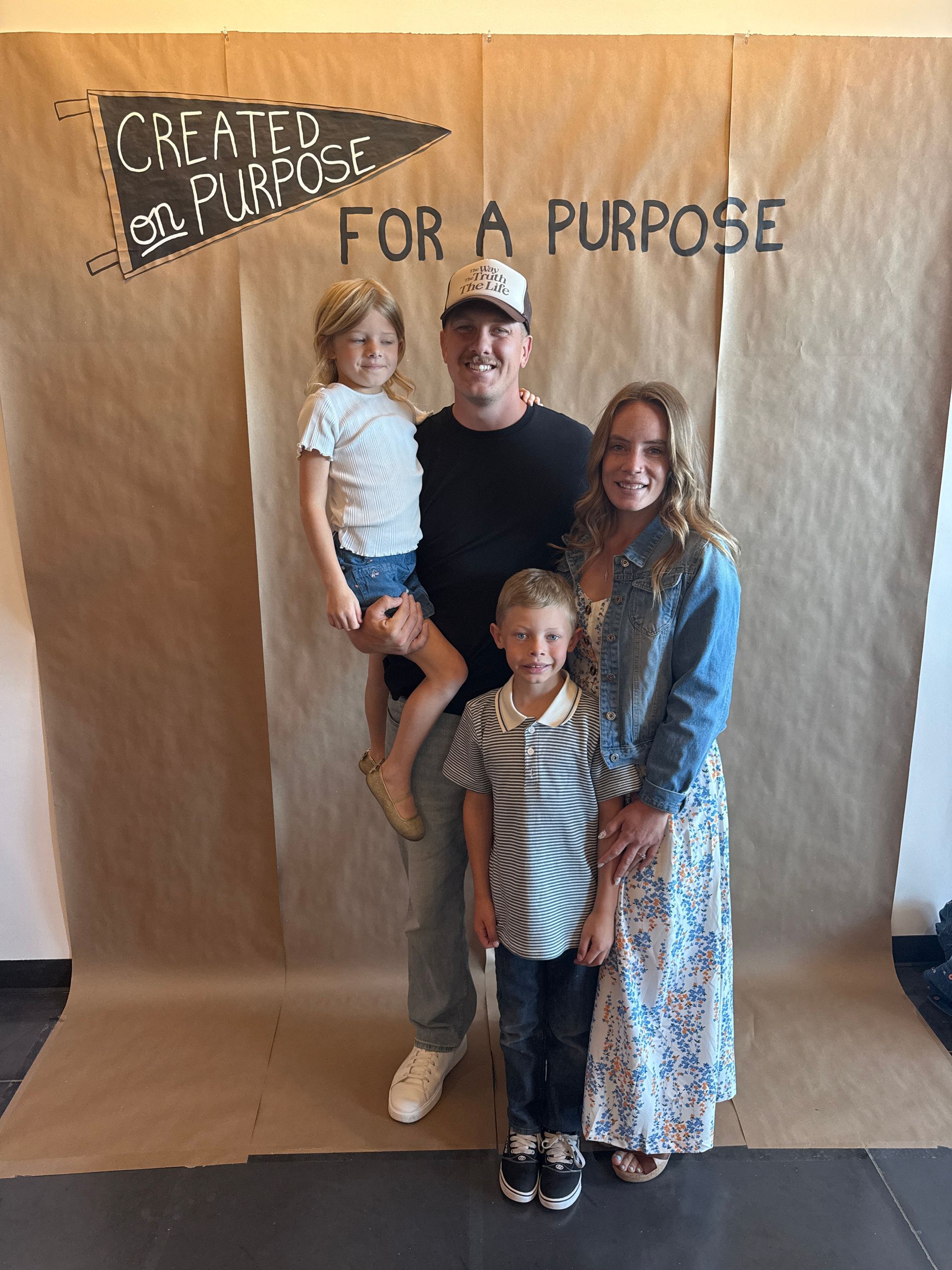 Family posing in front of brown paper backdrop with “Created for a Purpose” sign and banner.