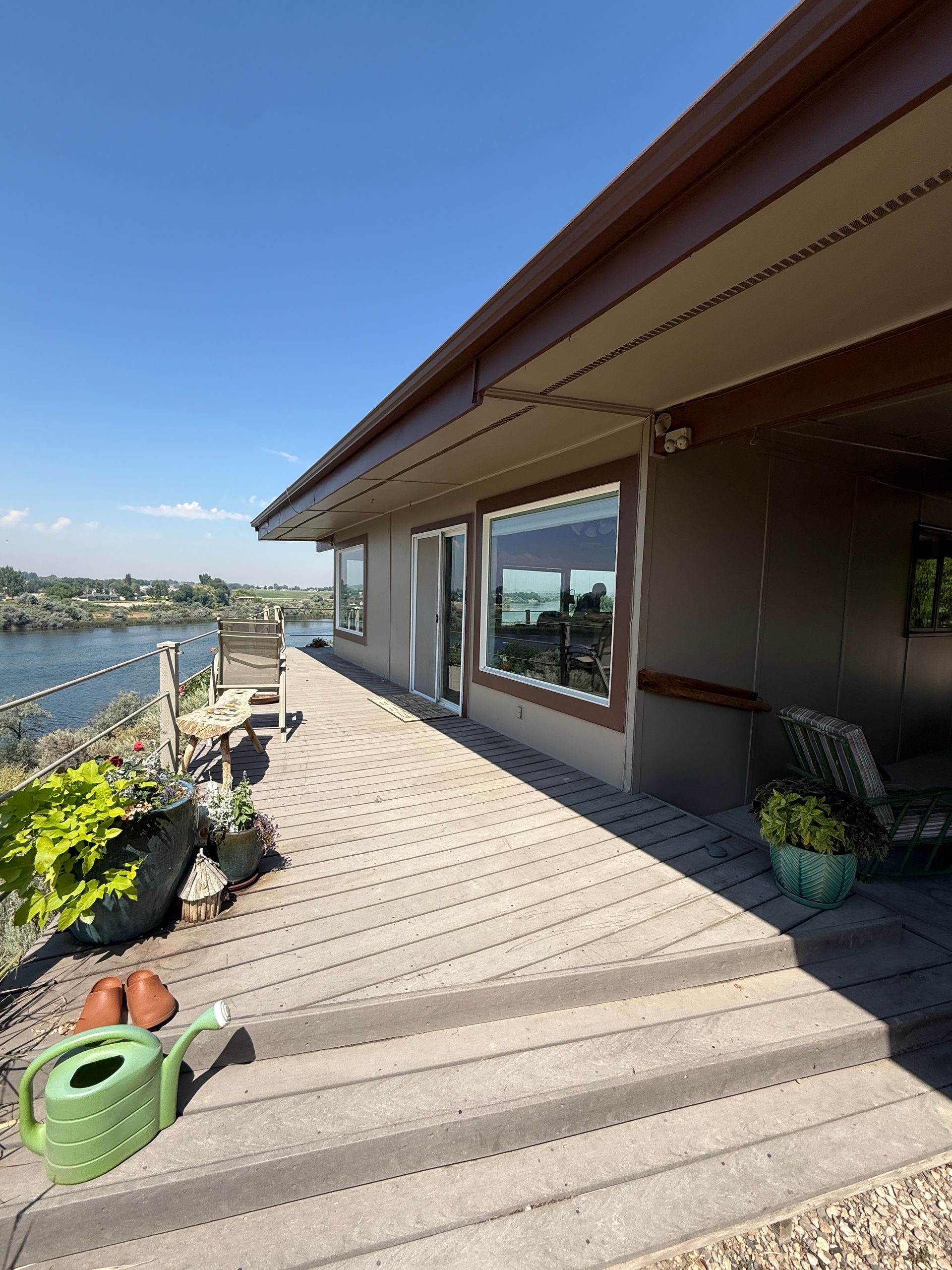 Riverside deck with potted plants, a green watering can, and a house window under clear blue sky