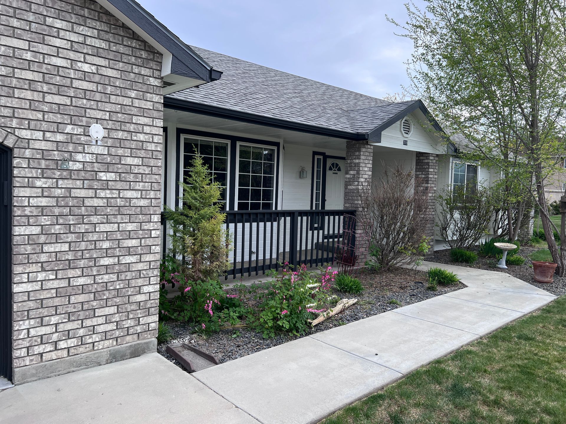 Brick house entrance with black porch railing, white trim, and a concrete walkway beside a flower bed