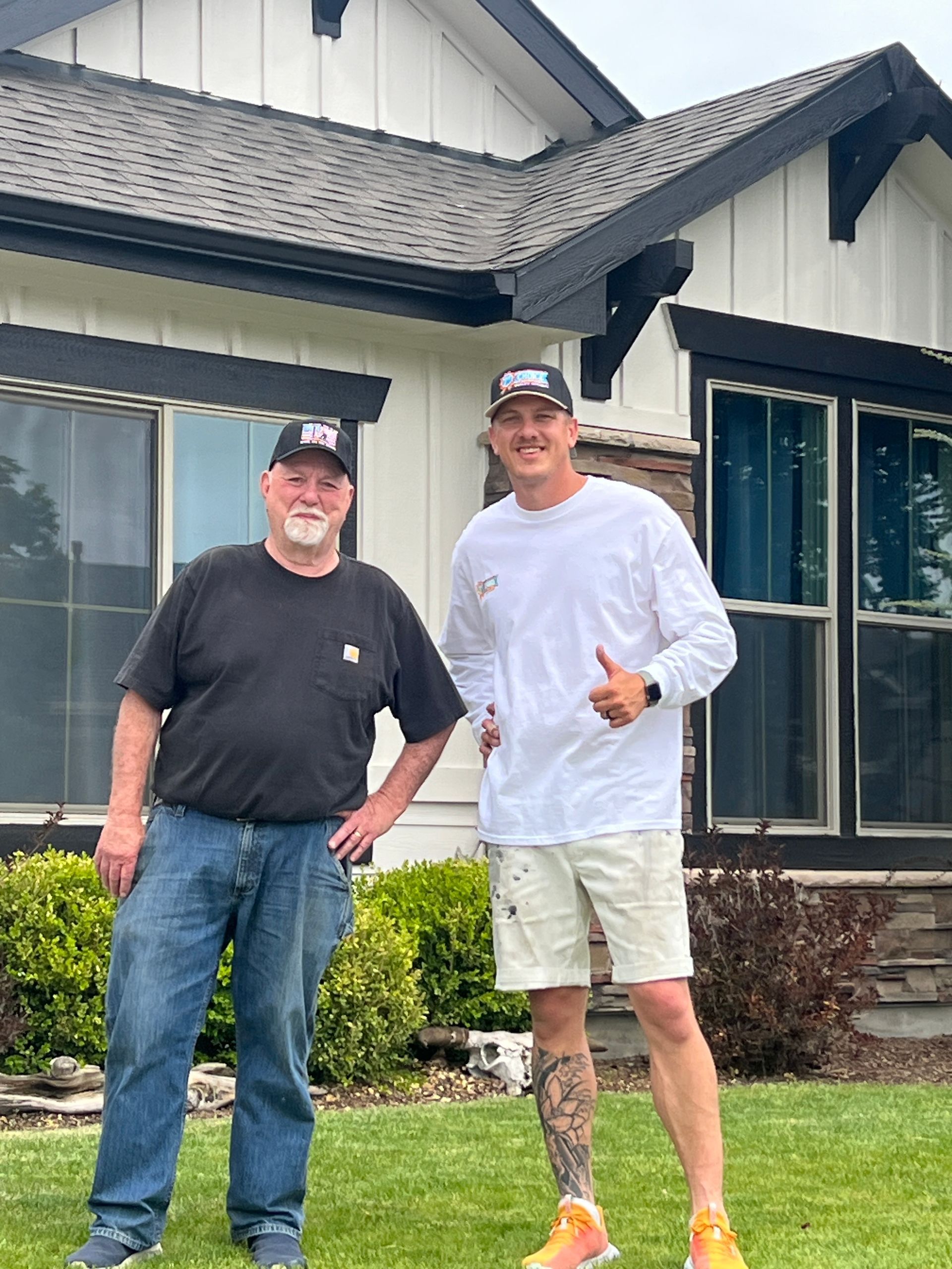 Two men standing and smiling outside a house, one giving a thumbs-up.
