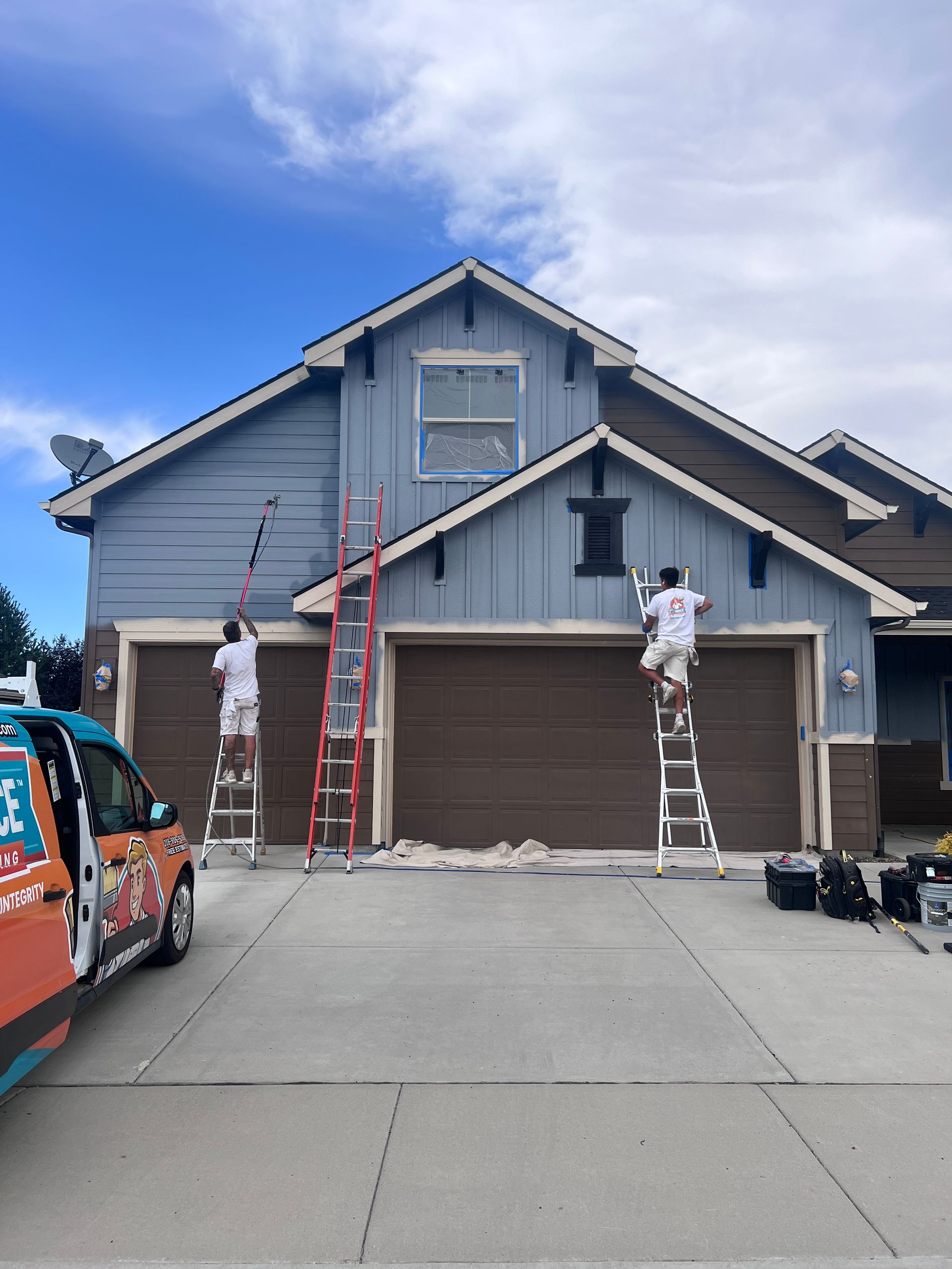 Two workers on ladders painting the front of a gray house with a brown garage door.