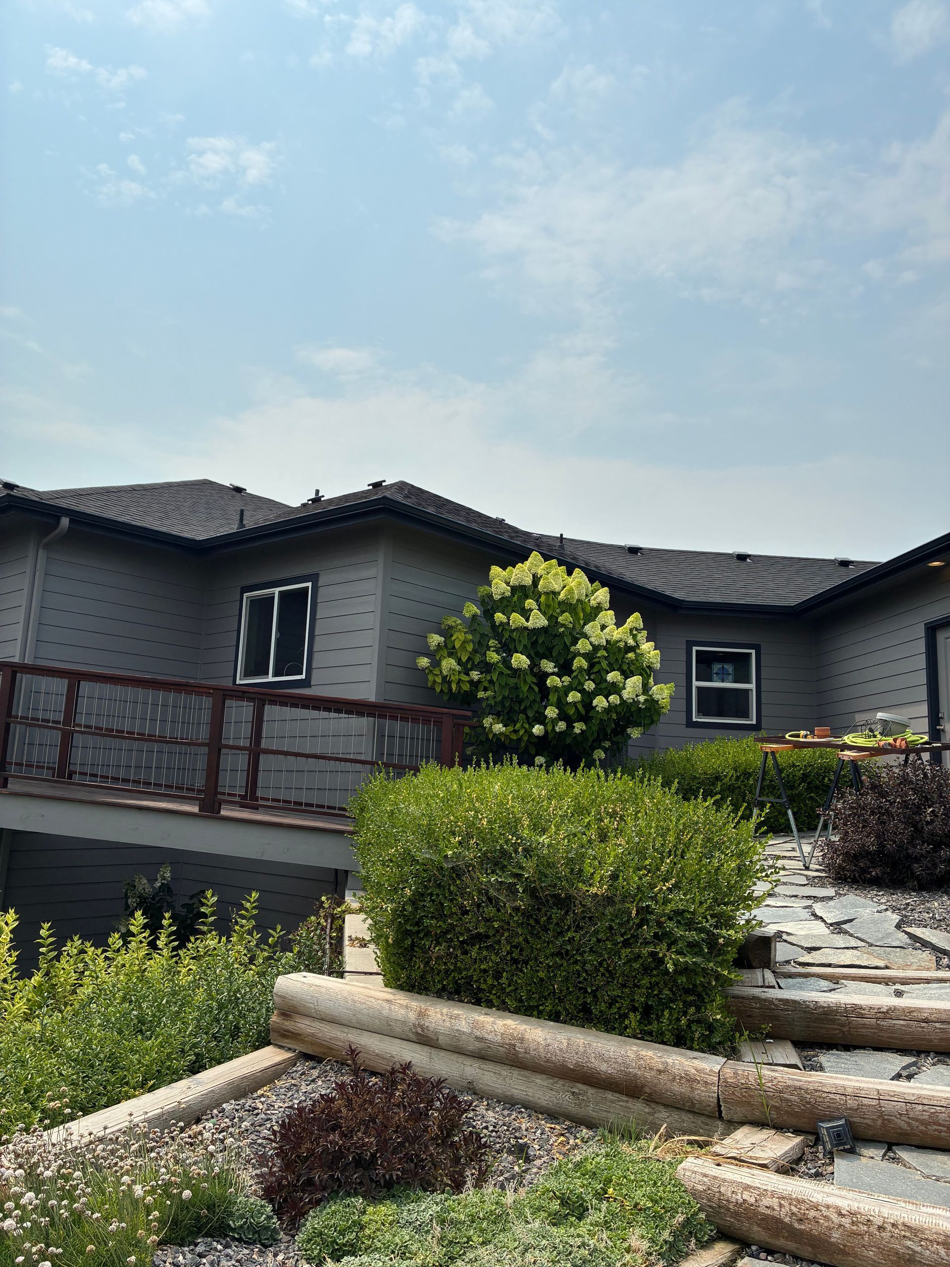 Modern gray house with dark roof, wooden deck, and landscaped front garden under a blue sky