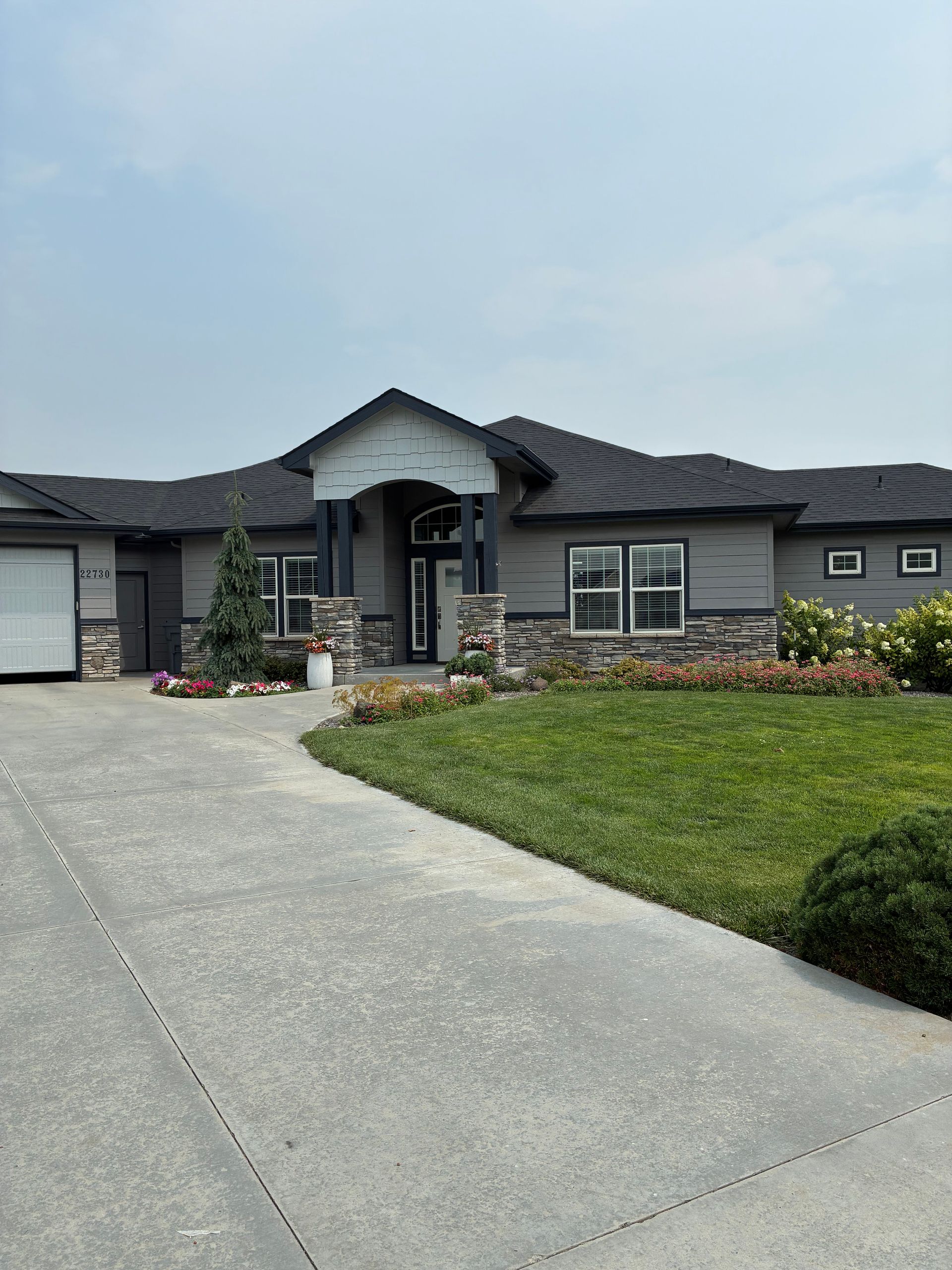 Single-story gray house with a curved driveway, front lawn, and flower beds under a cloudy sky
