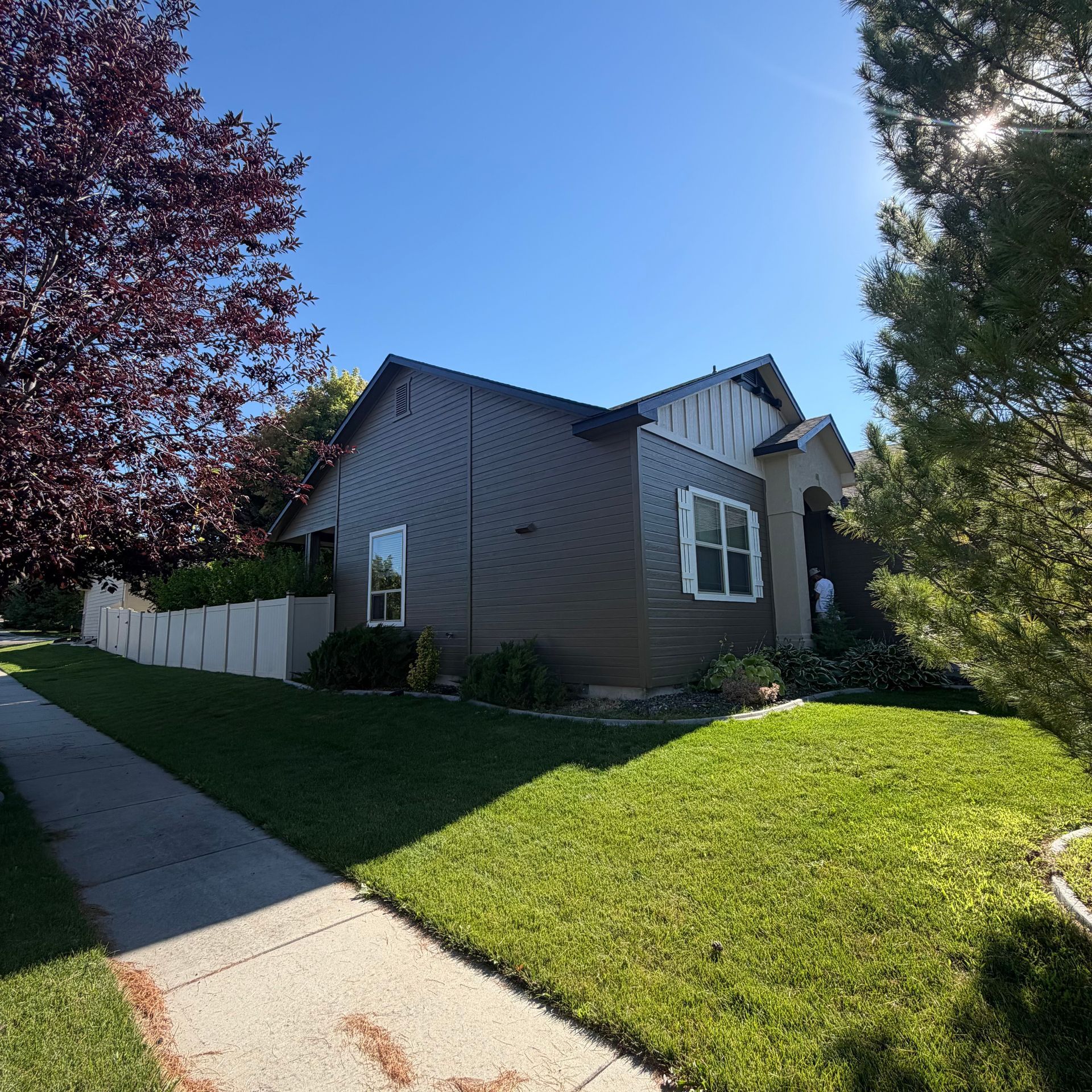 Suburban house with gray siding, green lawn, white fence, and sunny sidewalk