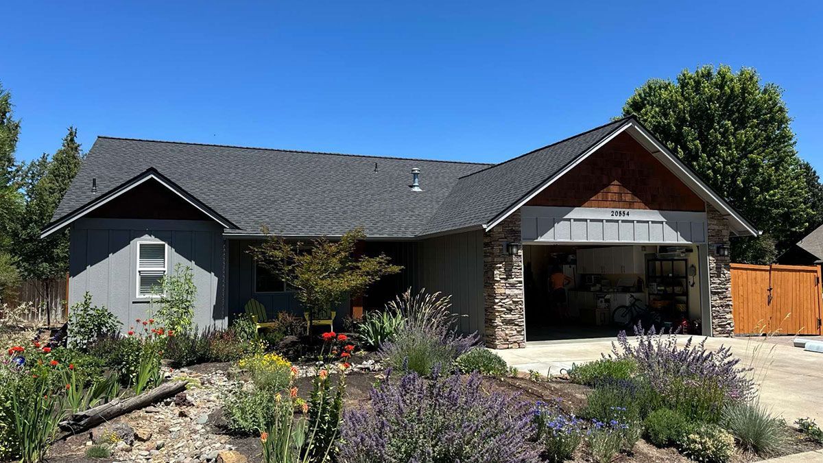 A house with a gray roof and a garage is surrounded by flowers and trees.