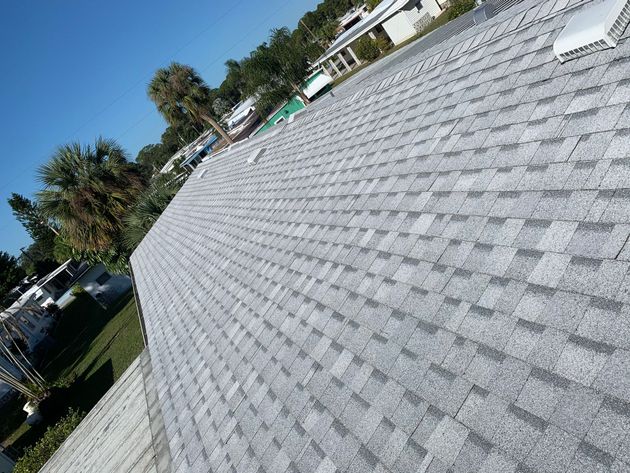 Tilted view of a gray shingle roof with trees and houses in the background under a clear sky