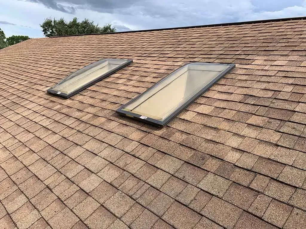 Roof with two rectangular skylights on brown shingles under a cloudy sky