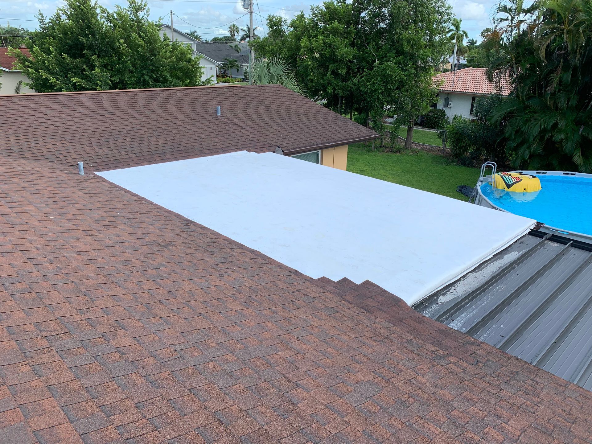 White tarp covering part of a brown shingled roof beside a gray metal roof and a blue pool