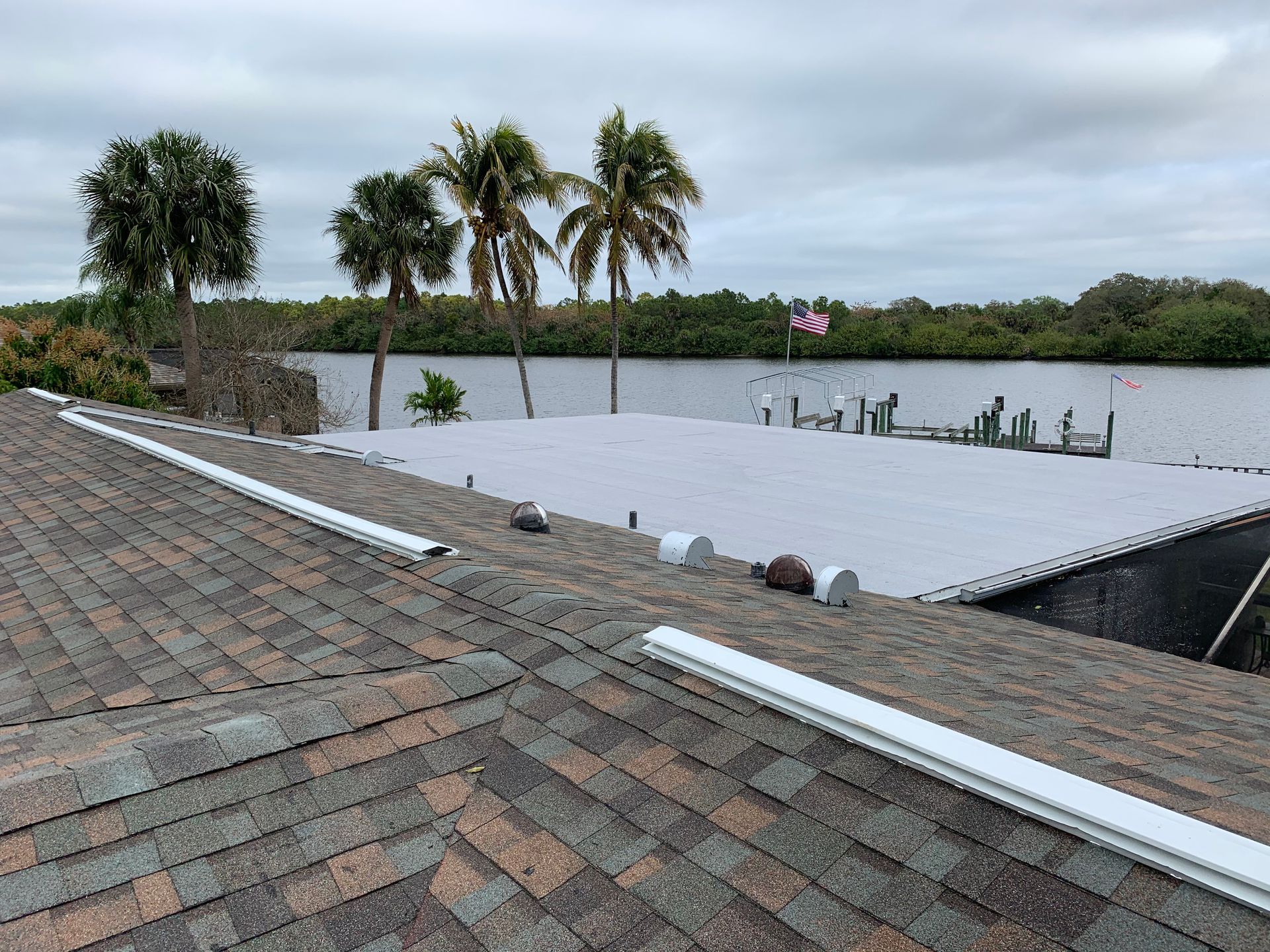 Rooftop with gray metal panels beside a waterway and palm trees under an overcast sky
