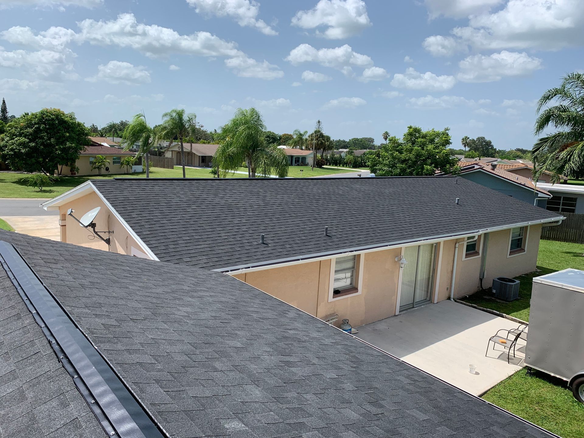 Aerial view of a single-story house with a gray shingle roof in a suburban neighborhood under a partly cloudy sky.