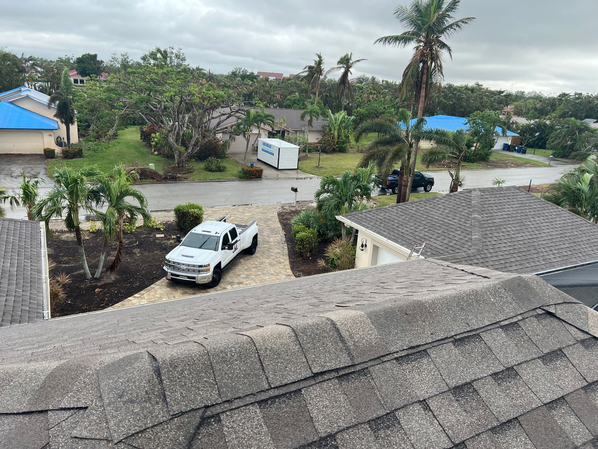 Flooded neighborhood street with a white pickup truck, palm trees, and gray rooftops under cloudy skies