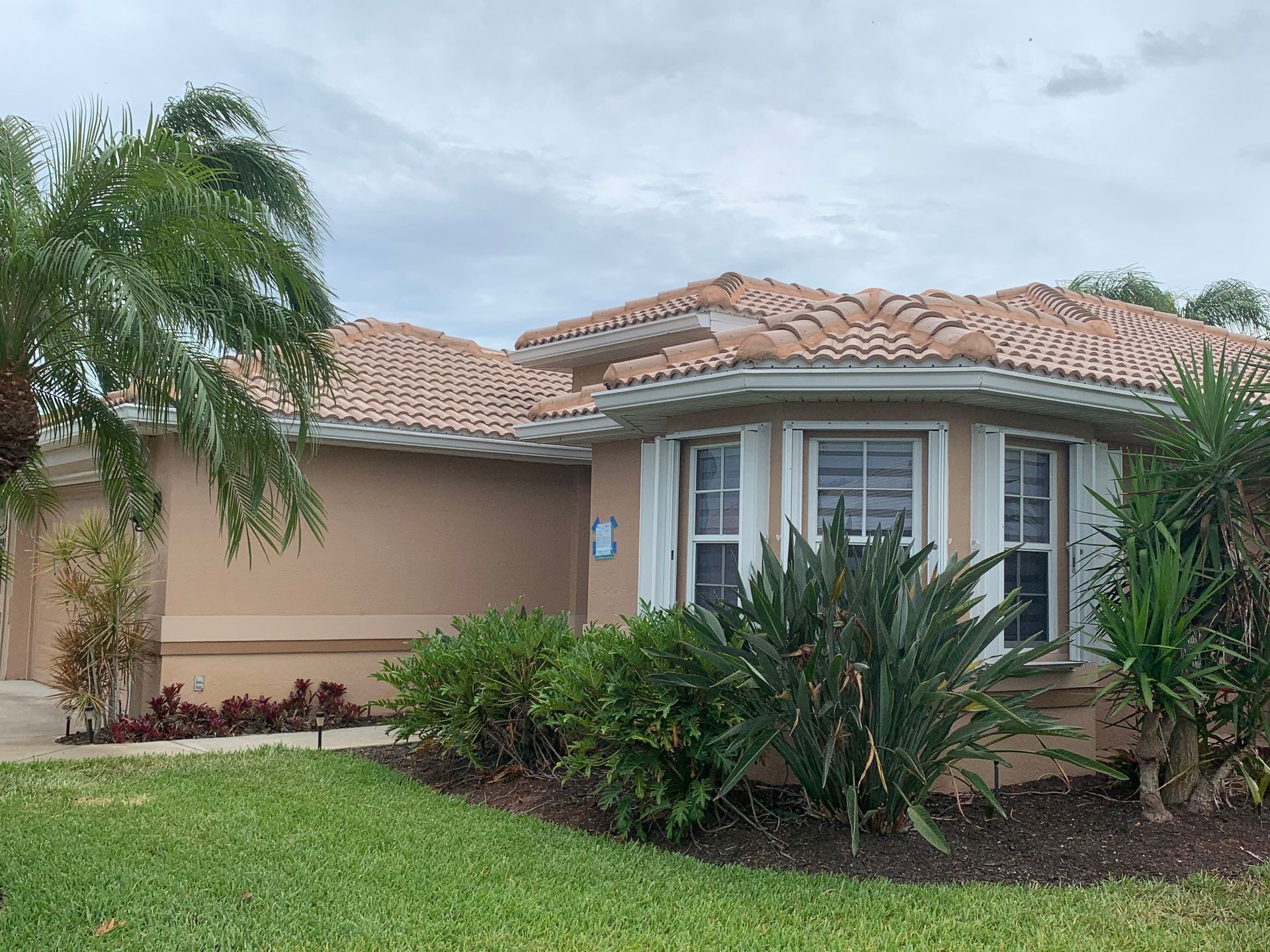 Single-story suburban house with tan stucco, terracotta roof, and lush front landscaping