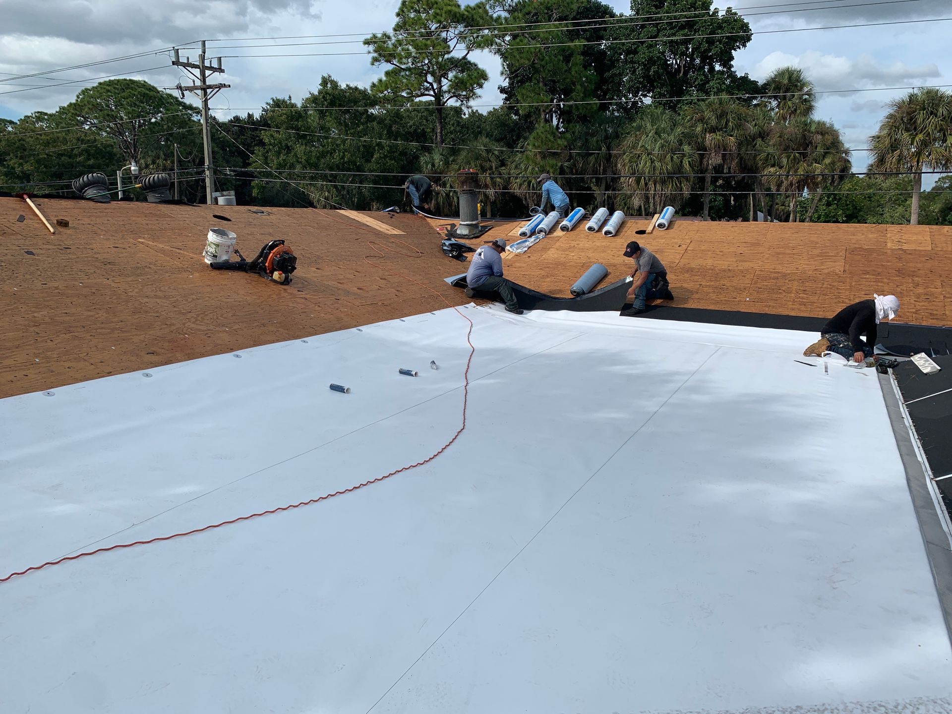 Workers installing a white rooftop membrane on a flat roof with a dirt embankment and trees nearby