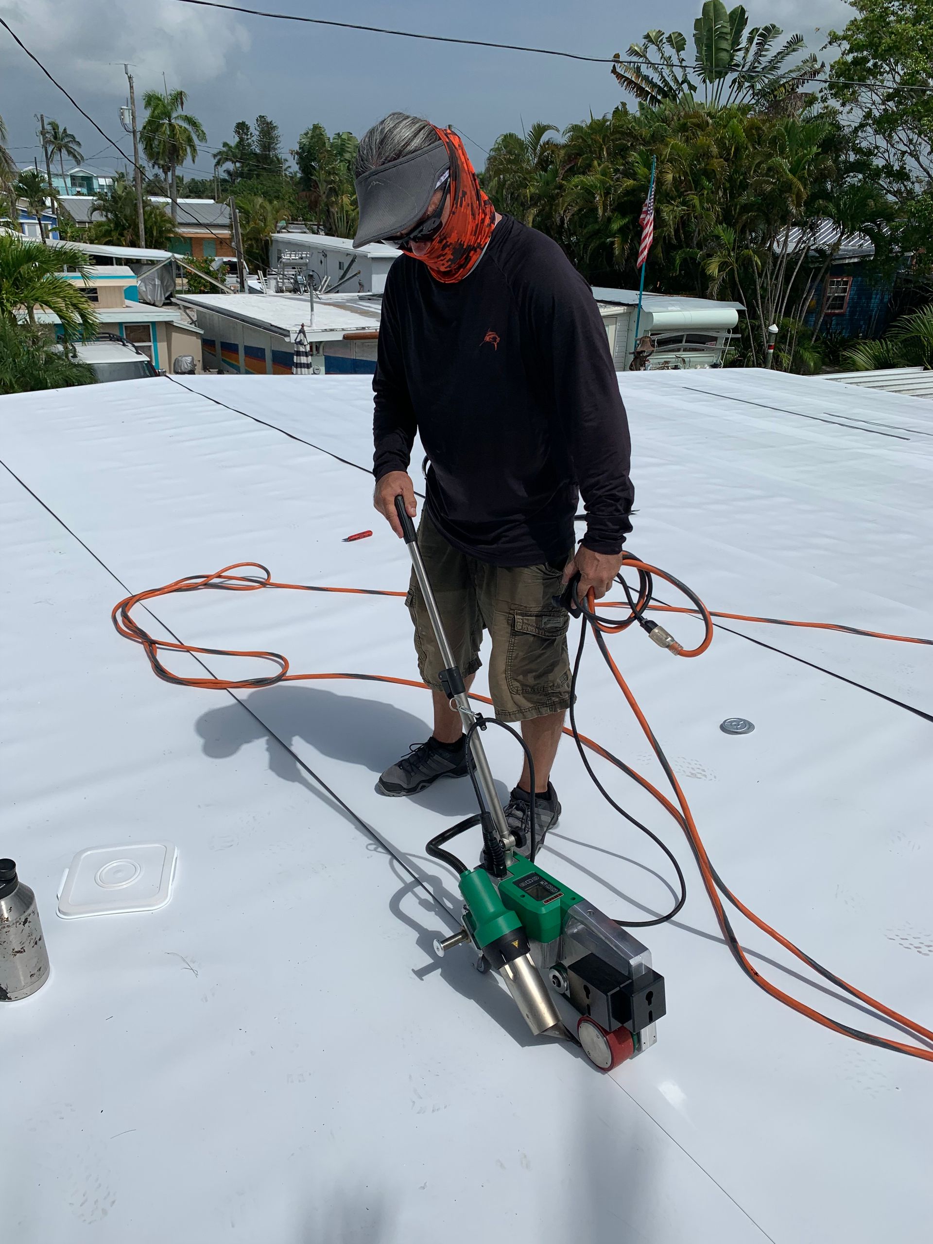 Worker on a white rooftop handling coiled orange cables near a green power tool, with trees and houses in the background.