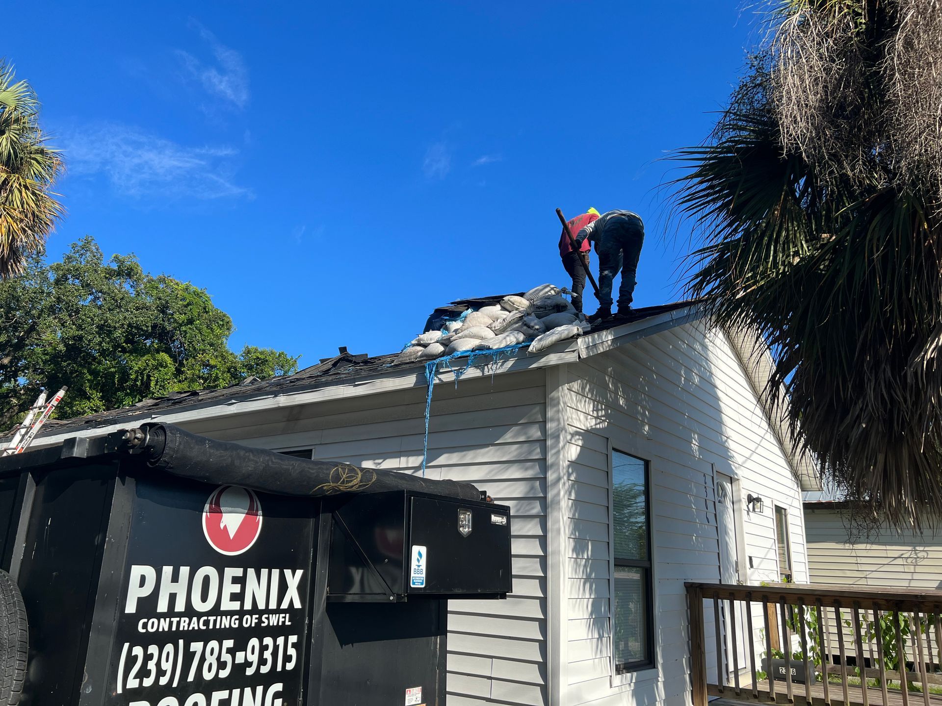 Workers on a roof above a Phoenix contractors sign, with a clear blue sky and trees nearby.