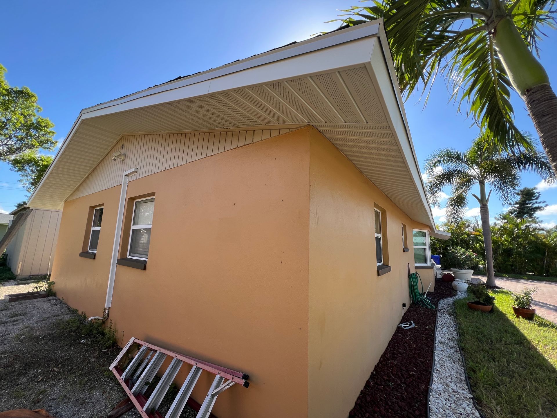 Tan stucco house exterior with white eaves, palm trees, and a ladder leaning against the wall
