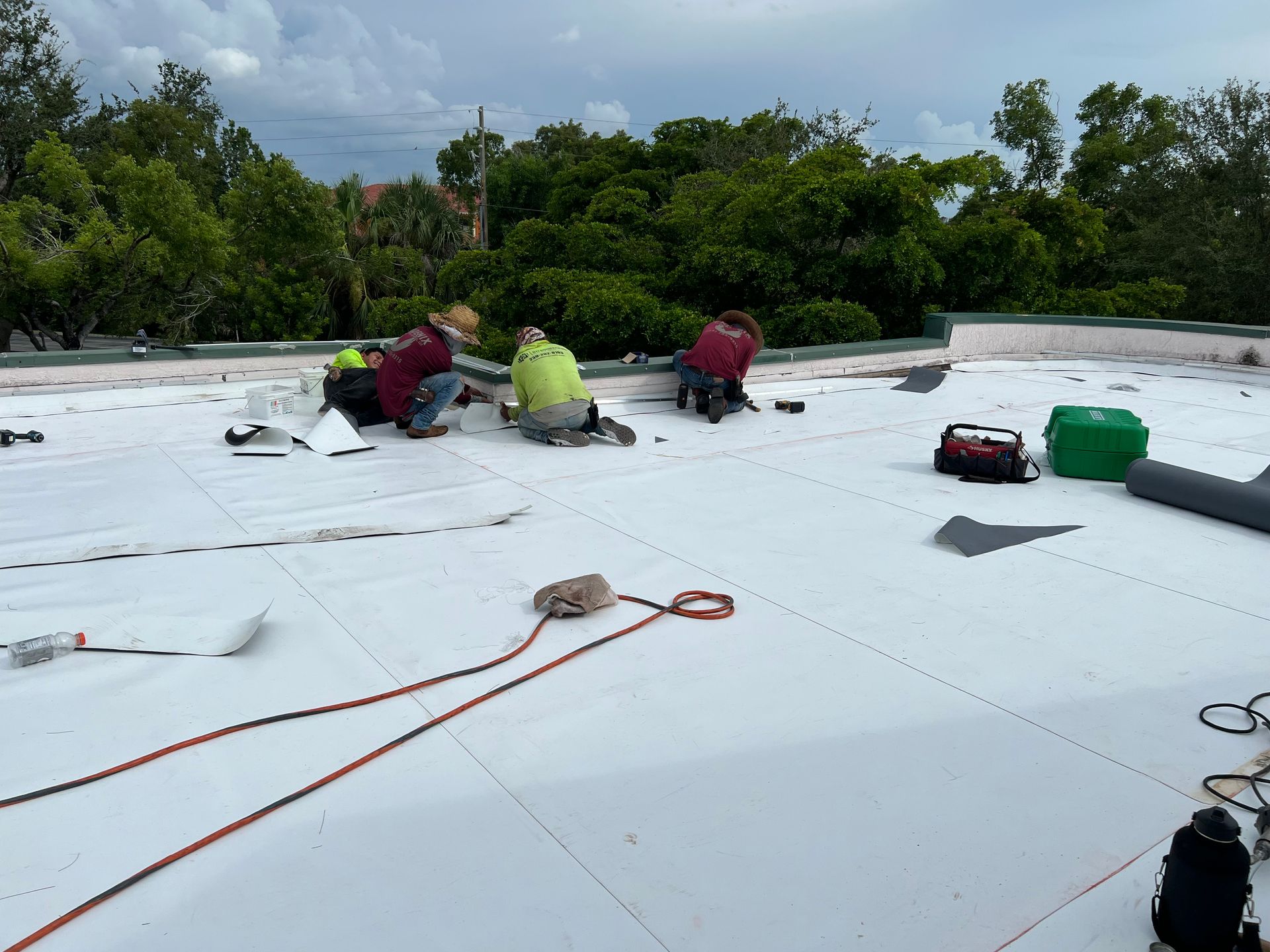 Workers on a white roof with tools and orange rope, surrounded by trees under cloudy sky
