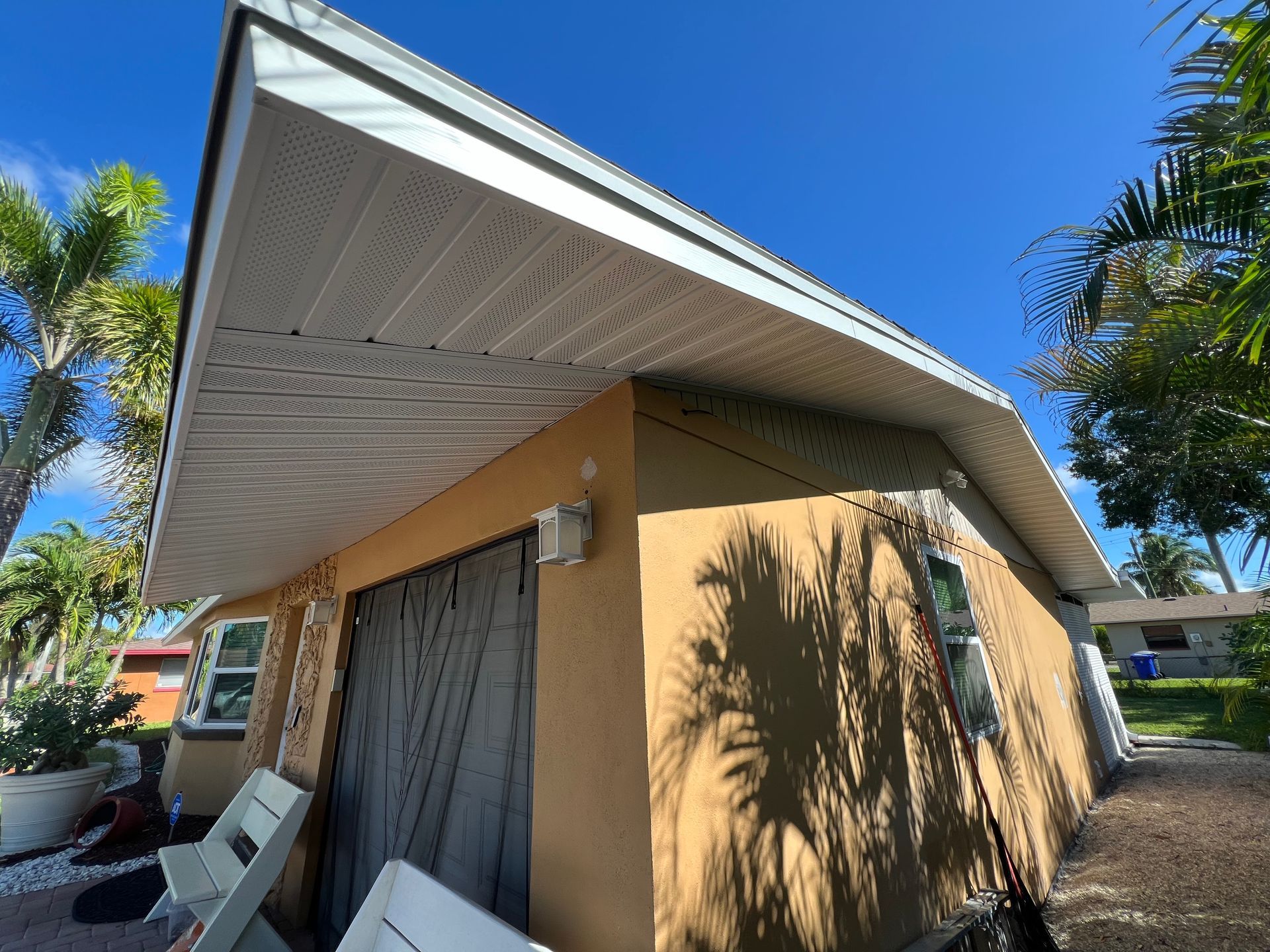 Tan house with steep white roof, patio curtains, and palm trees under a bright blue sky