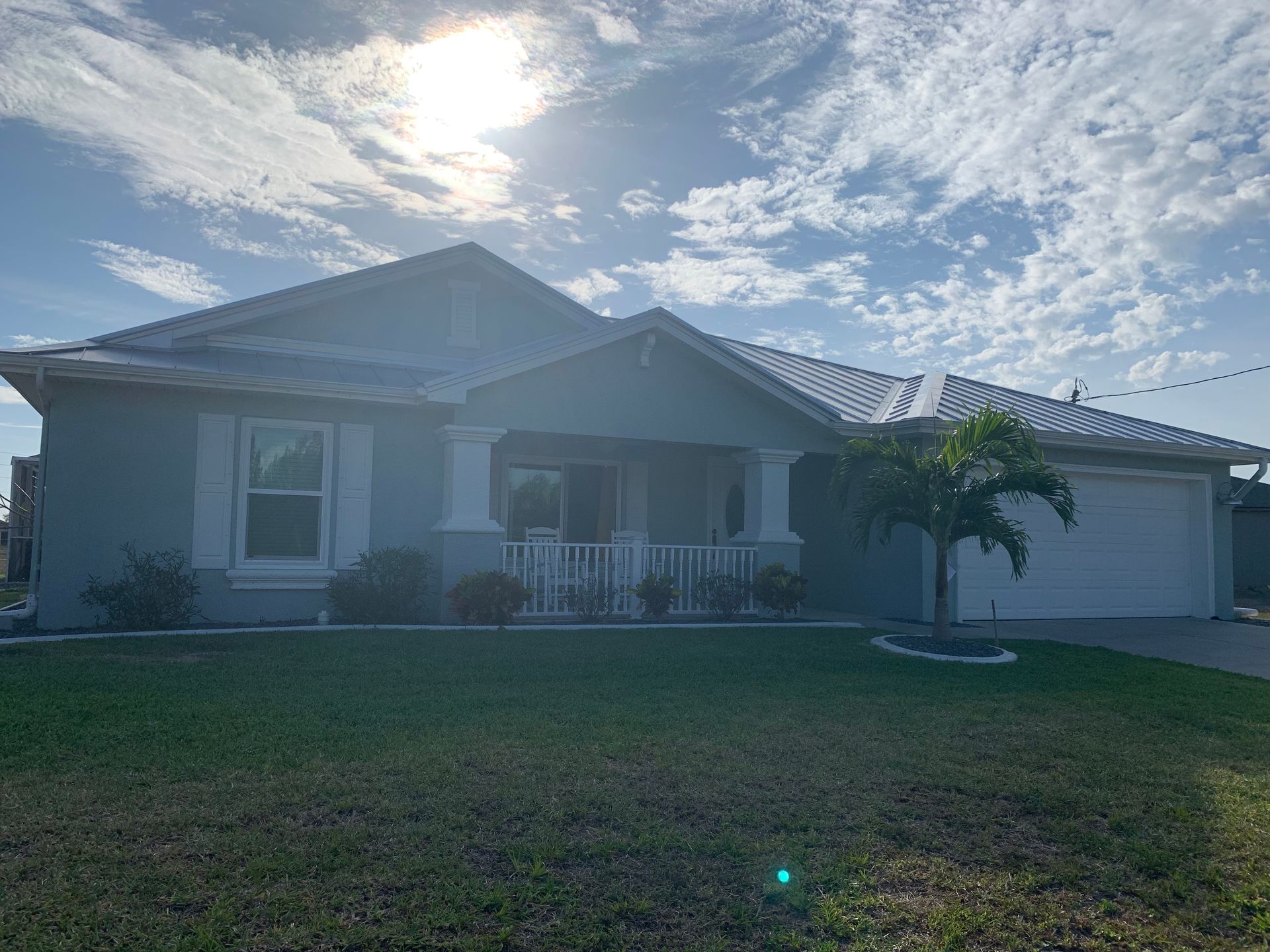 Single-story house with a front lawn, garage, and a small palm tree under a bright cloudy sky.