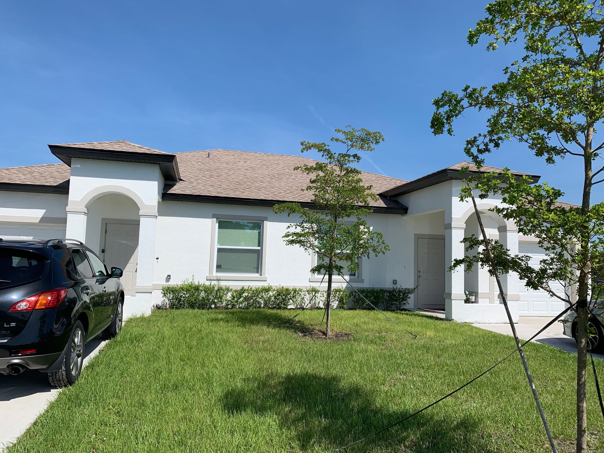 White house with a tiled roof, front lawn, trees, and a parked black car under a blue sky