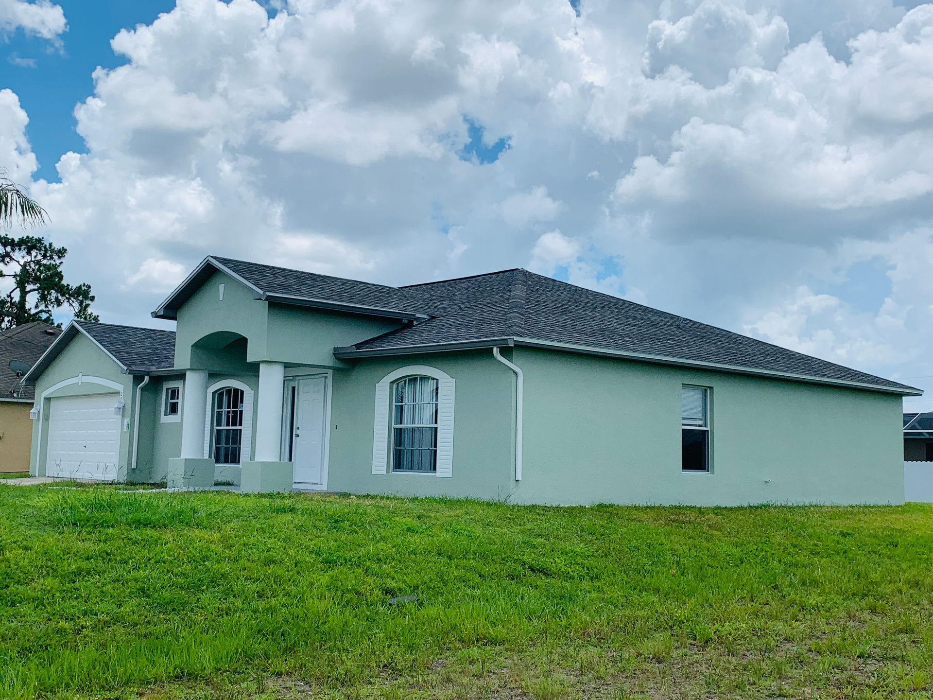 Single-story pale green house with dark roof in a grassy field under a cloudy sky