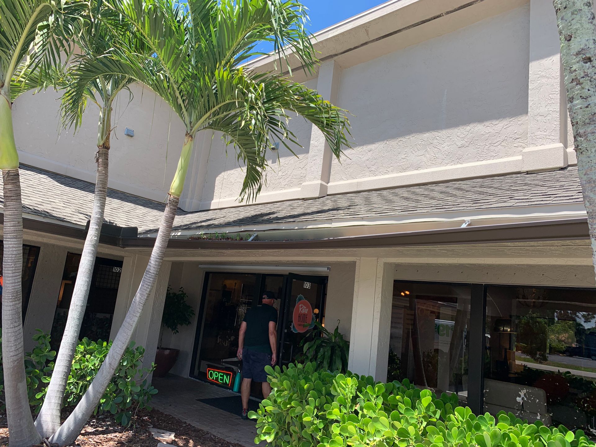 Palm trees shade a resort-style building with a covered entrance and people standing near the doorway.