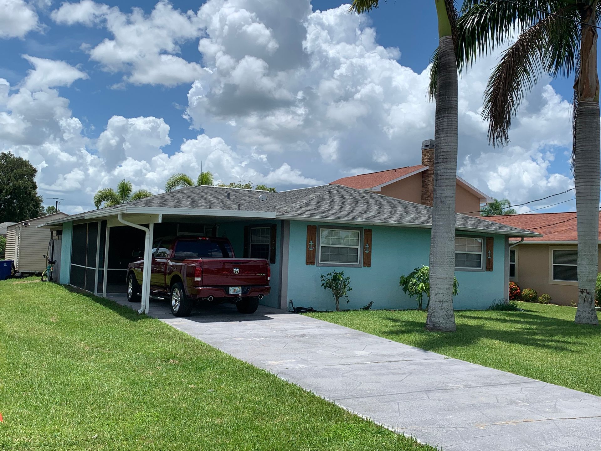 Single-story suburban house with a red truck in the driveway, a carport, palm trees, and a blue sky.