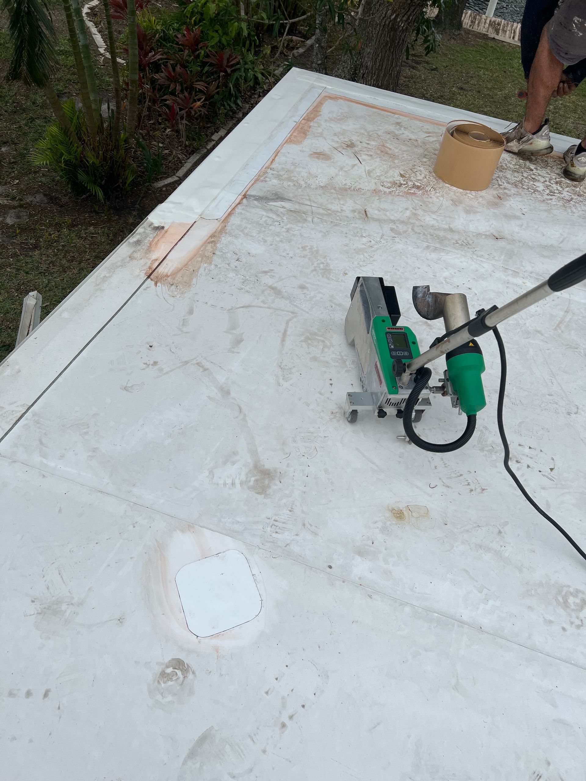 White rooftop with a green power tool and hose near a person’s legs, beside a small bucket and roof patches