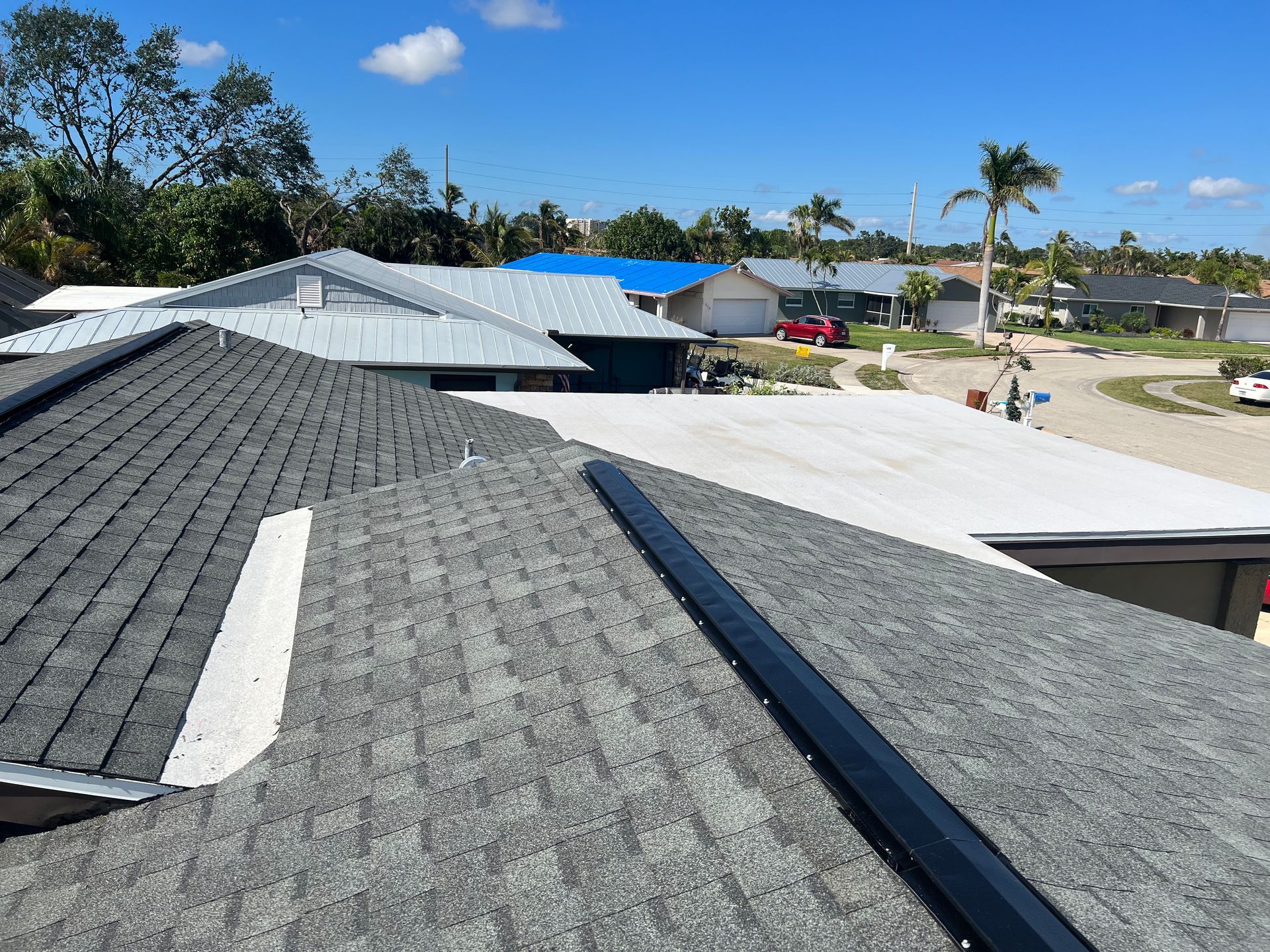 Rooftop view of gray and white shingle roofs in a sunny suburban neighborhood with blue sky and palm trees