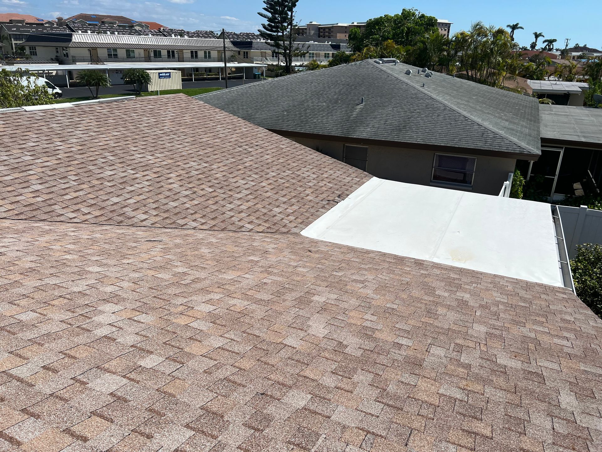 Residential rooftops with shingle roofs, a white flat roof, and distant houses under a clear sky