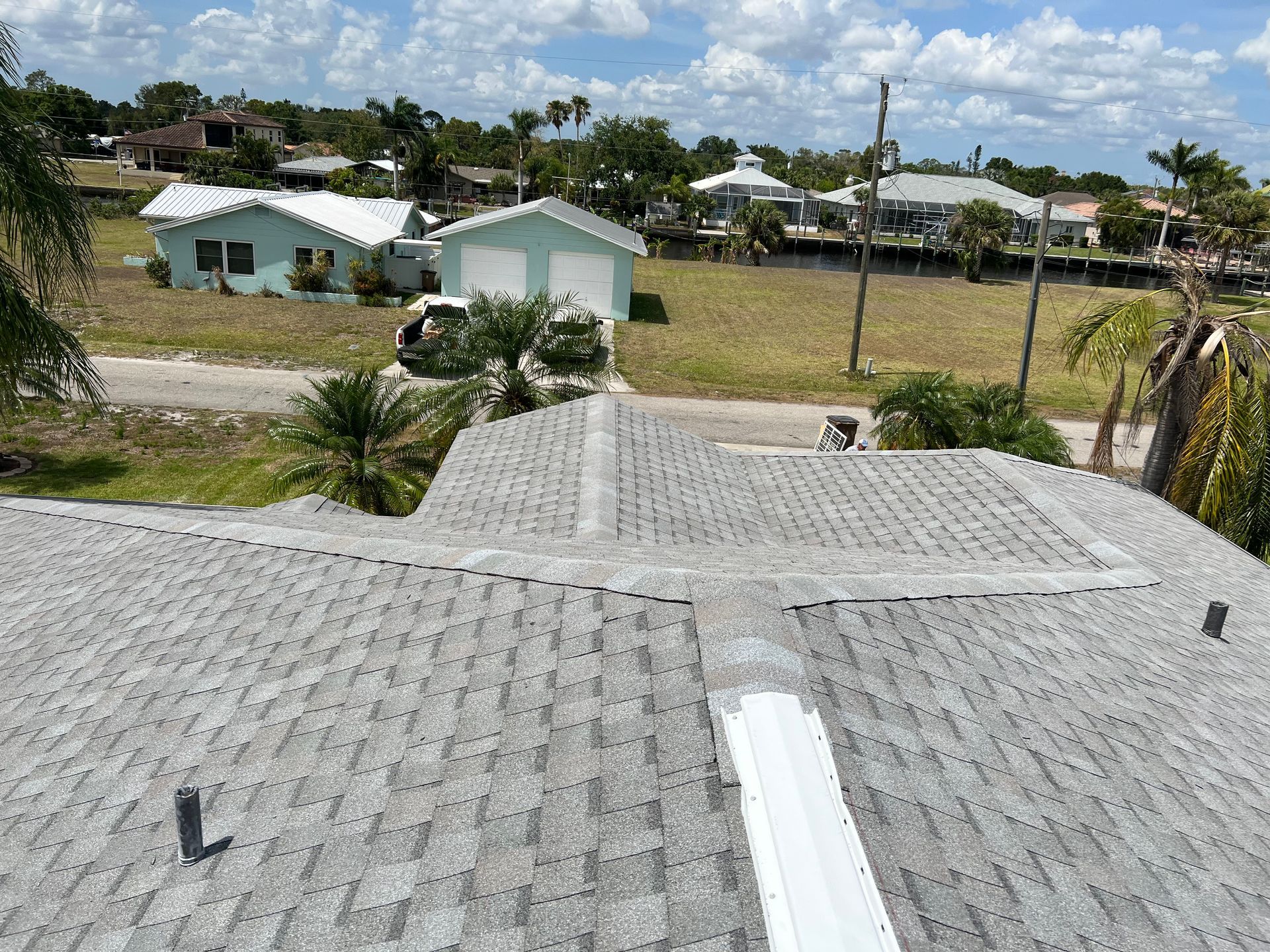 View from a shingled rooftop over a suburban neighborhood with small houses, palm trees, and a cloudy sky