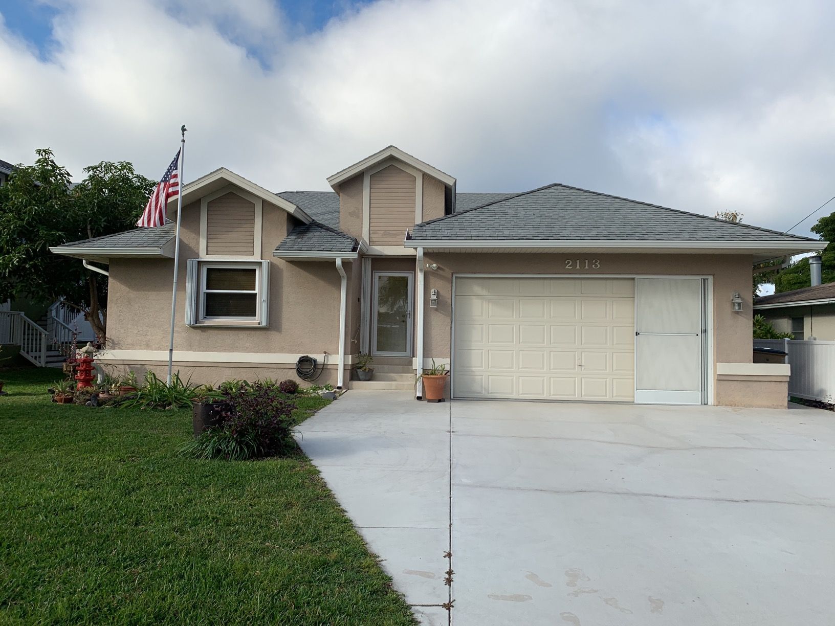 Suburban beige house with a two-car garage, American flag, and wide driveway under a cloudy sky