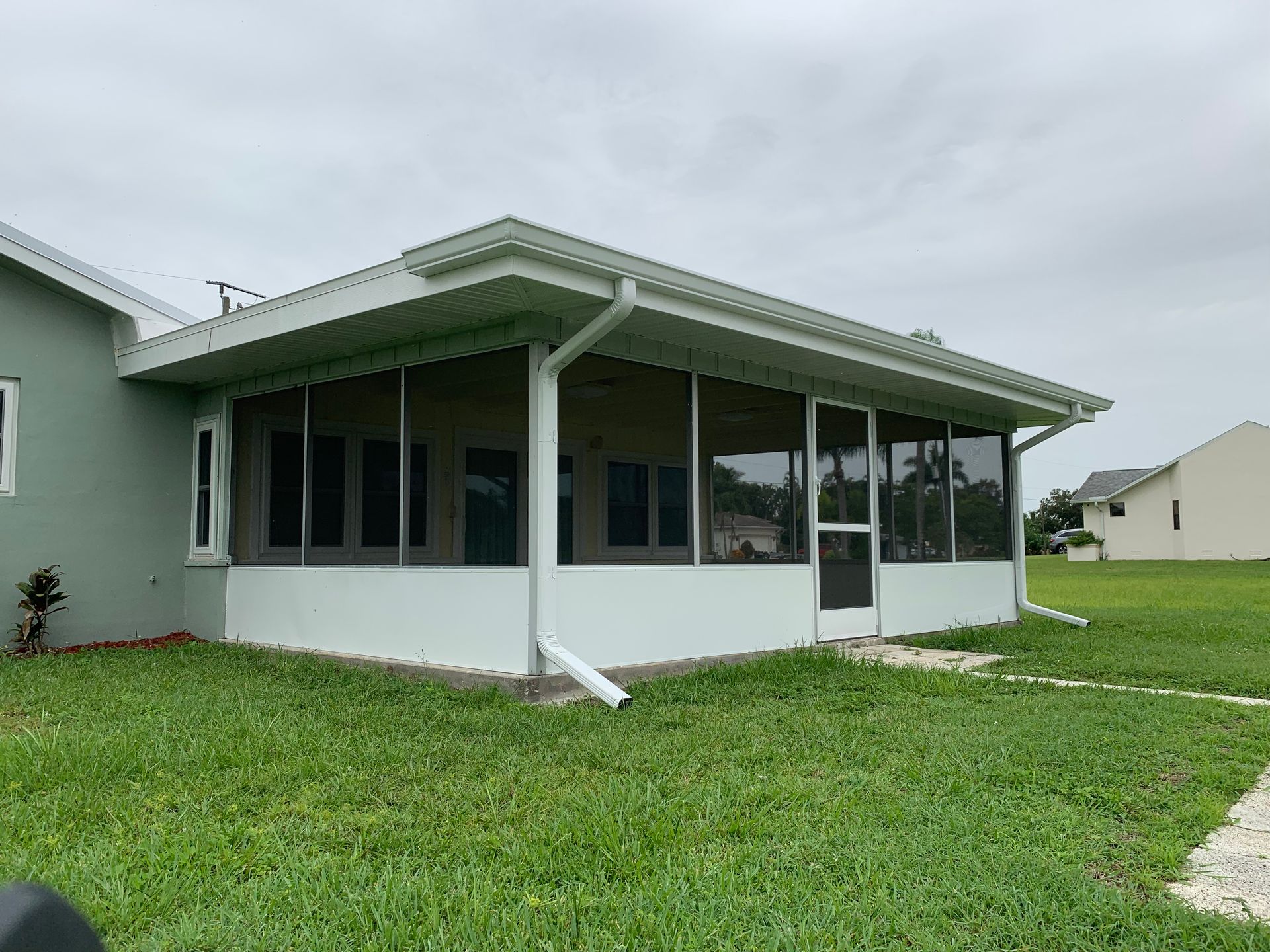 Single-story light green house with screened porch and white trim on a grassy lot under cloudy skies