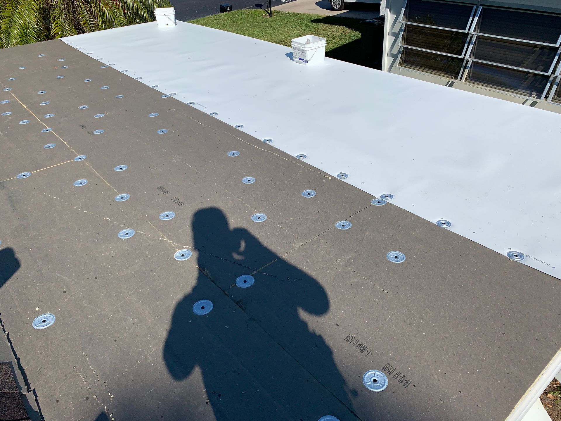 Flat rooftop with white and brown sections, dotted with fasteners; a person’s shadow is visible in the foreground.