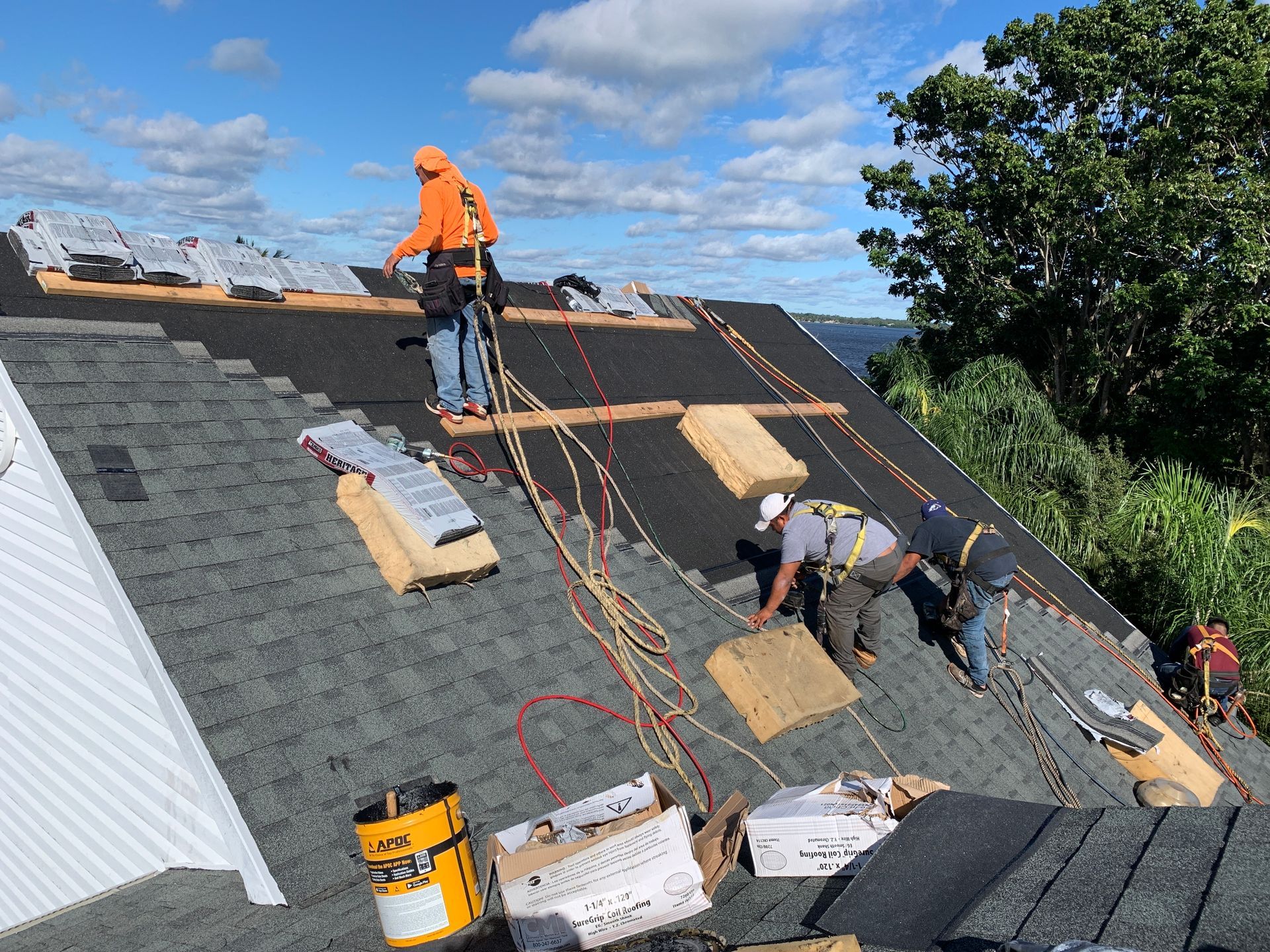 Workers installing roofing shingles on a steep roof under a blue sky.
