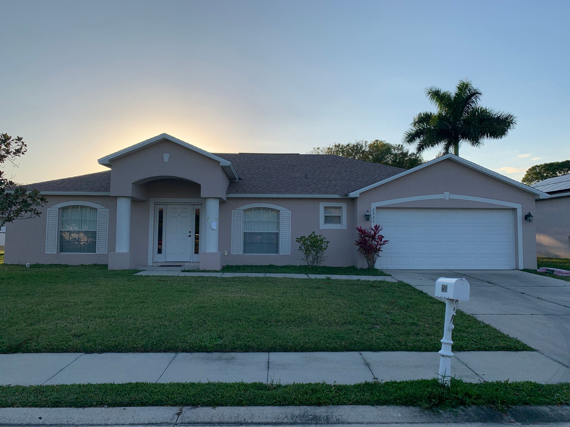 Single-story suburban house with garage, light pink facade, and a sunset sky over the front lawn