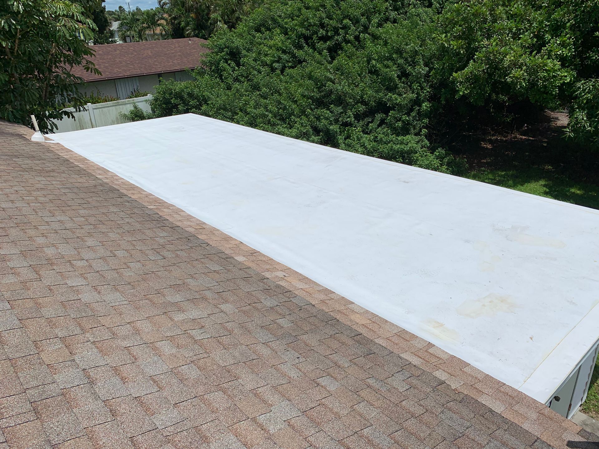 White flat rooftop beside a tan shingled roof, with trees and a house in the background