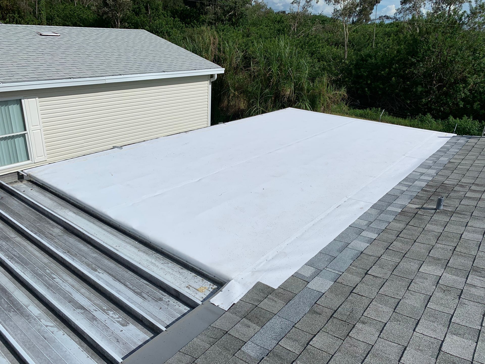 White roof tarp secured over a flat roof beside gray shingles and a shed.