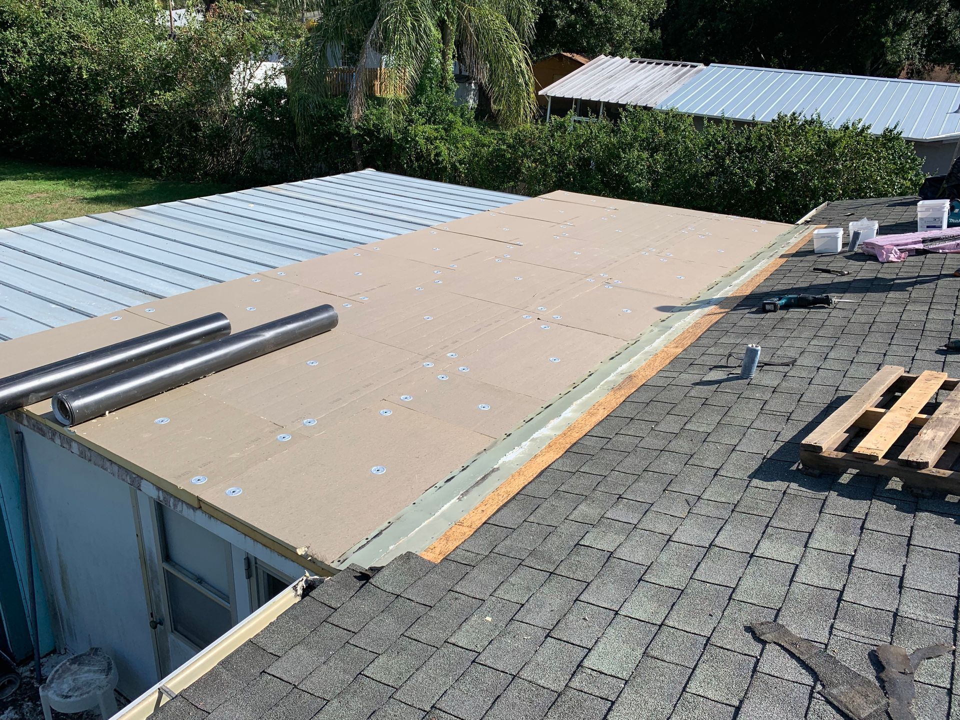 Rooftop construction with plywood sheathing, exposed beams, and stacks of roofing materials on a sloped roof.