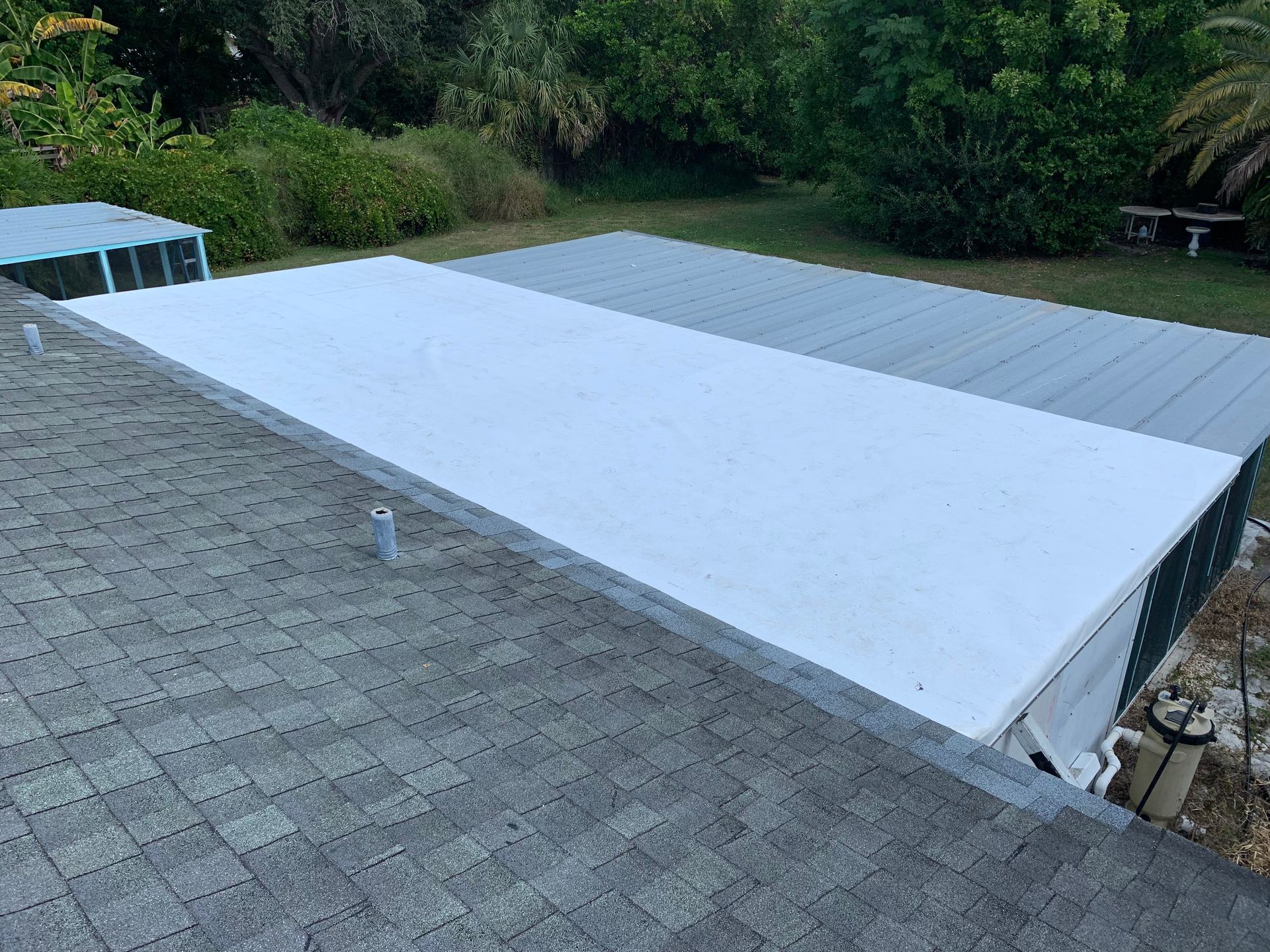 Rooftop with gray shingles and a large white tarp-covered section beside a wooded yard