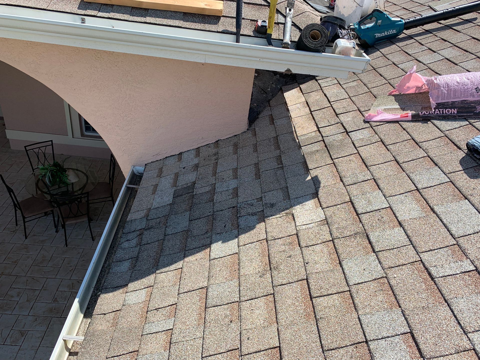 Roof shingles being repaired beside a white house with a shaded patio below.