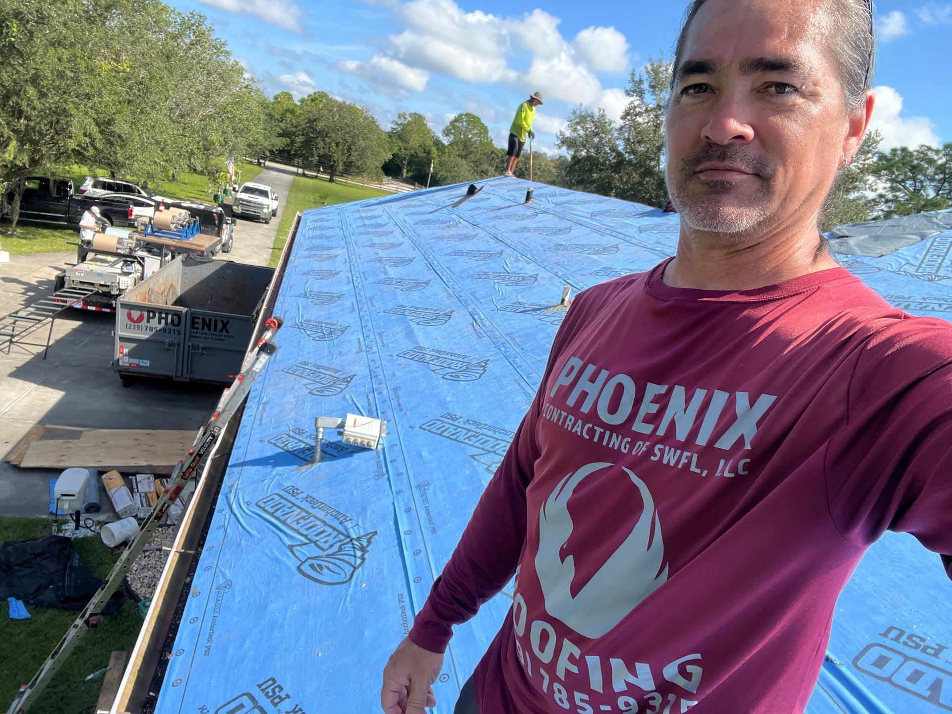Worker on a blue roof under construction, wearing a maroon Phoenix Roofing shirt.