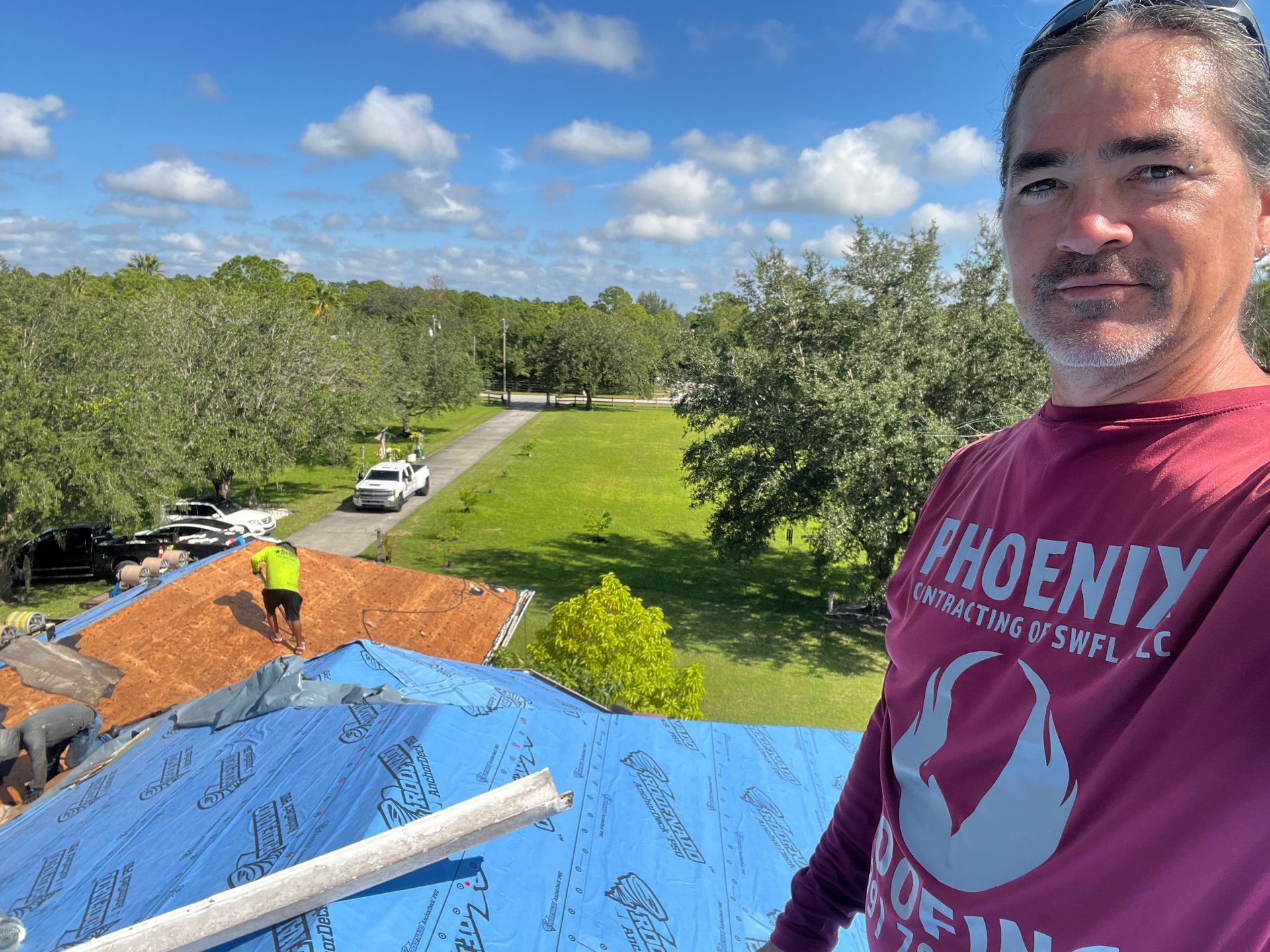 Person on a roof taking a selfie while another worker stands by a blue tarp, with trees and cars below.