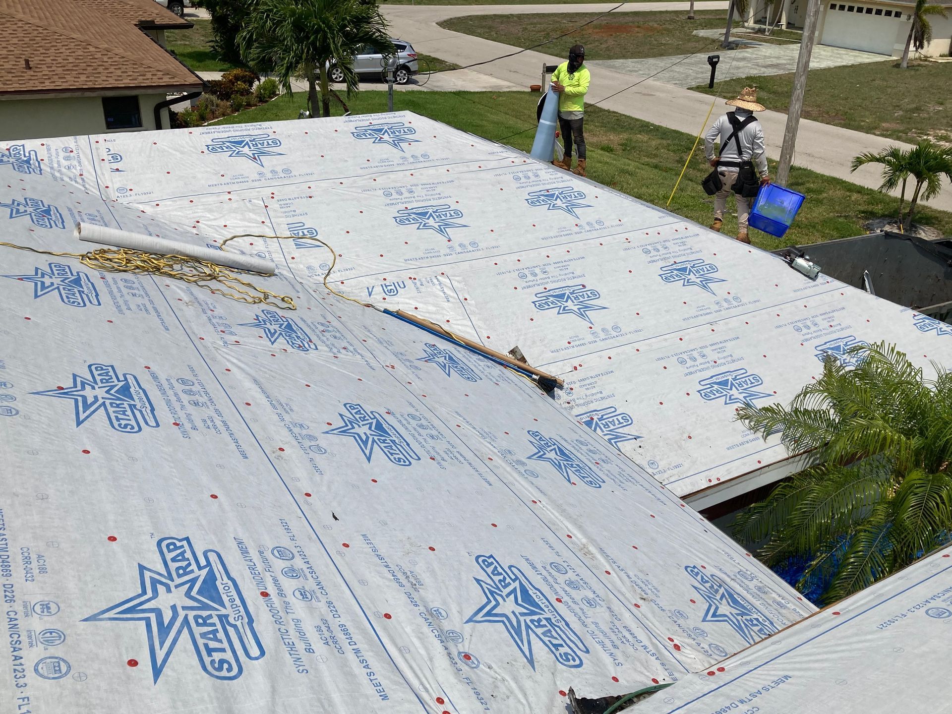 Workers installing white roofing underlayment on a roof, with tools and debris nearby.