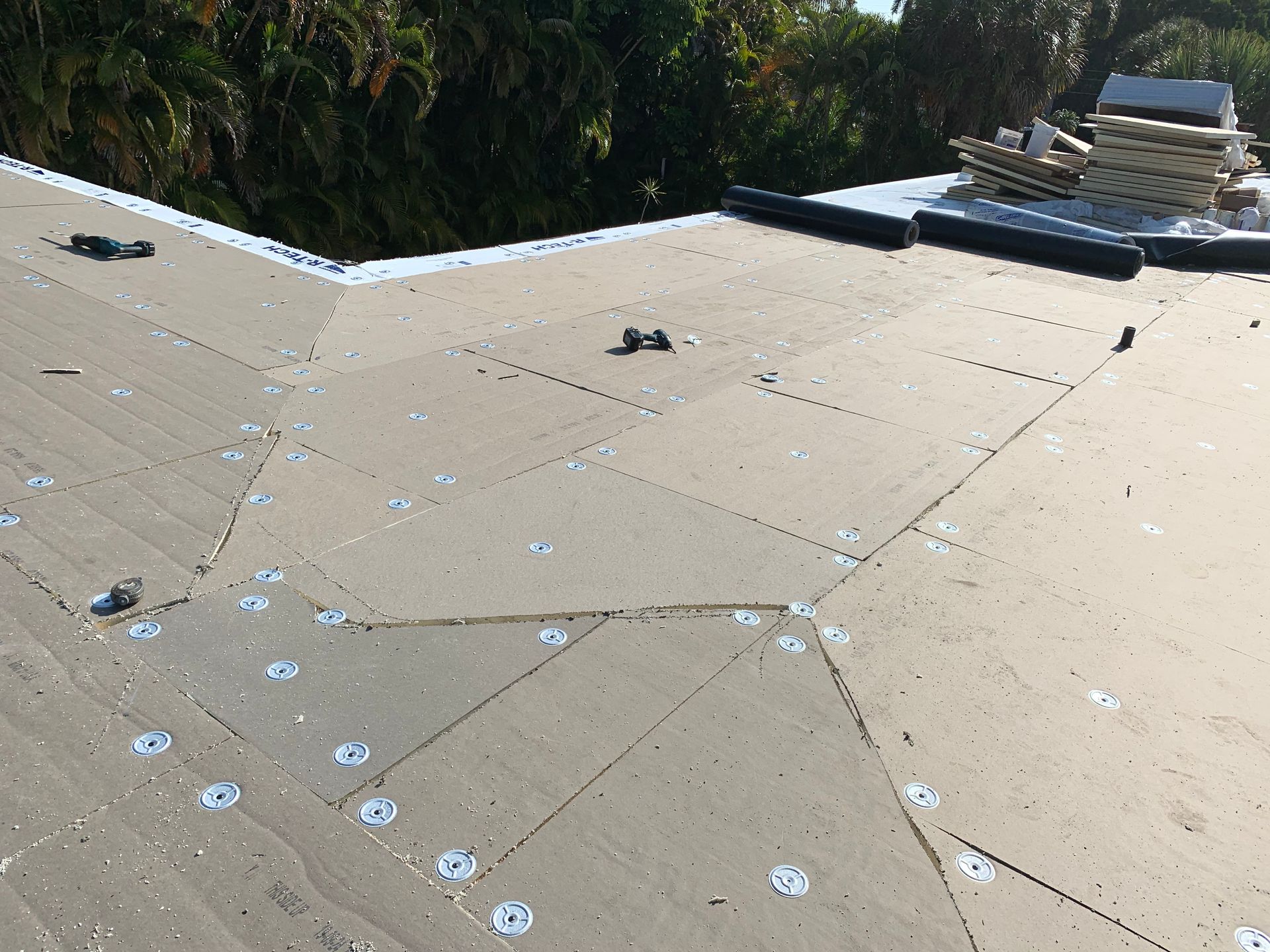 Flat tan rooftop with seams and scattered fasteners, bordered by trees and a small structure in the distance