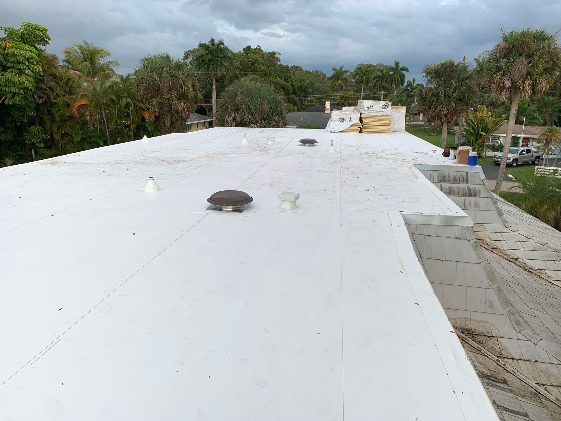 White flat roof with vents and equipment, bordered by palm trees and cloudy sky.