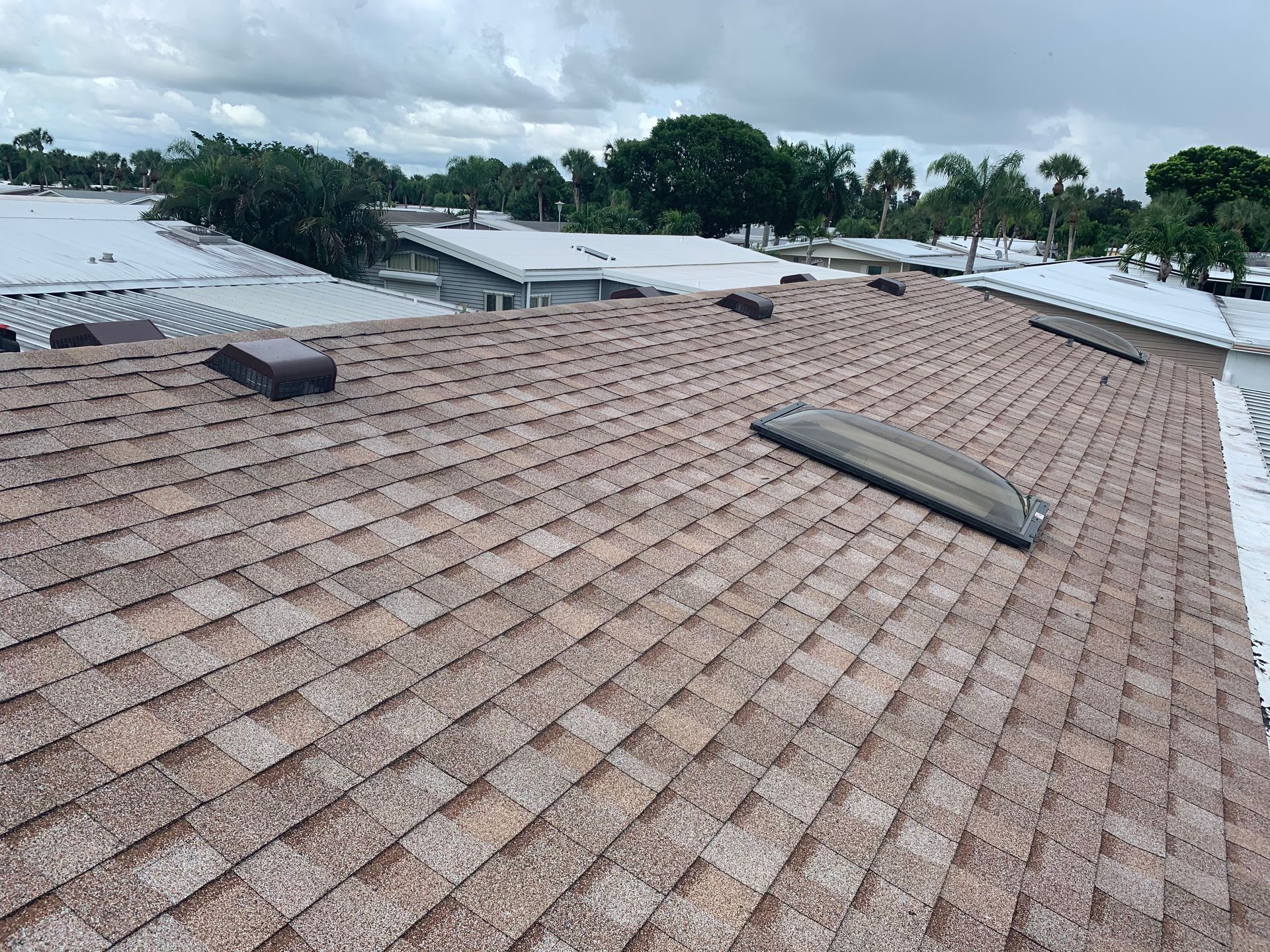 Rooftop covered with brown shingles, damaged ridge vent, and stormy sky over nearby homes.