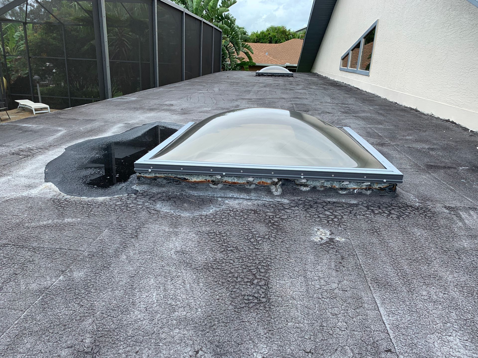 Flat skylight partially covered by white tarp on a gravel rooftop beside a black fence and white building